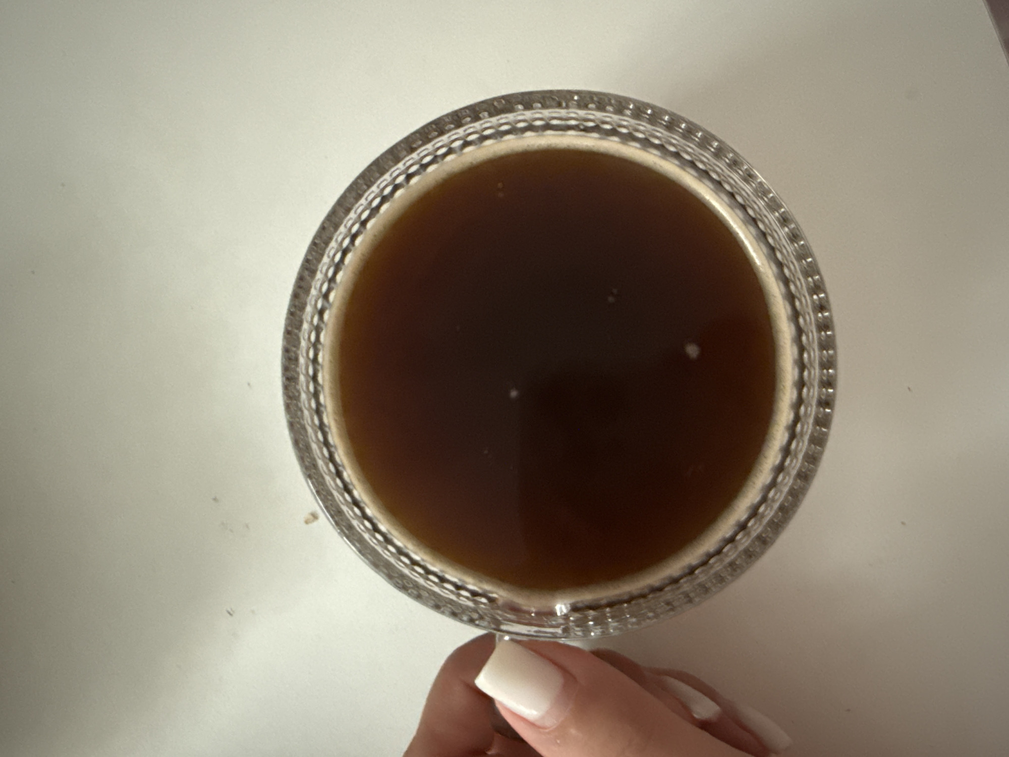 A close-up, top-down view shows a clear glass mug filled with dark brown liquid, likely coffee, set against a plain white surface. The mug's rim has a decorative textured pattern. A person's hand with neatly manicured white fingernails is visible on the left side of the frame, delicately holding the mug's handle. The surface appears to be a table or countertop, devoid of any other objects or distractions, suggesting a simple and focused scene. There are no discernible features that would place this image in a specific city or country, including Vologda, Russia.