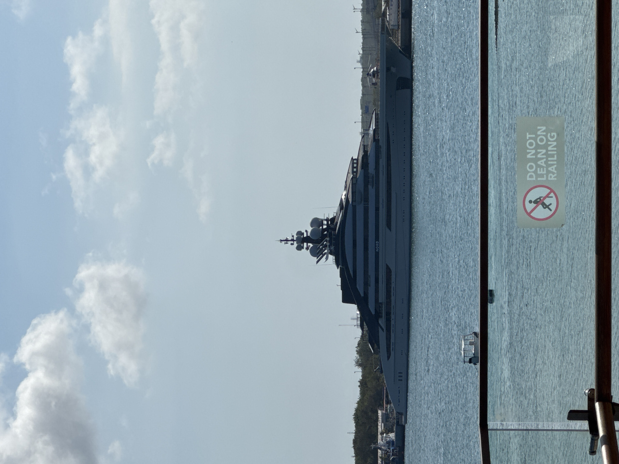The image shows a large, multi-deck superyacht docked in a harbor. The yacht is light-colored with dark accents and has a sleek, modern design. It is equipped with multiple radar domes and antennas on its upper decks. The yacht has the name "NORD" visible on its side. In the background, there is a treeline and industrial structures, suggesting a port or coastal city. A smaller boat is visible in the water near the yacht, and a sign with a prohibition symbol and the text "DO NOT LEAN ON RAILING" is in the foreground, indicating the viewpoint is from another vessel or balcony. The sky is partly cloudy, and the water is calm.