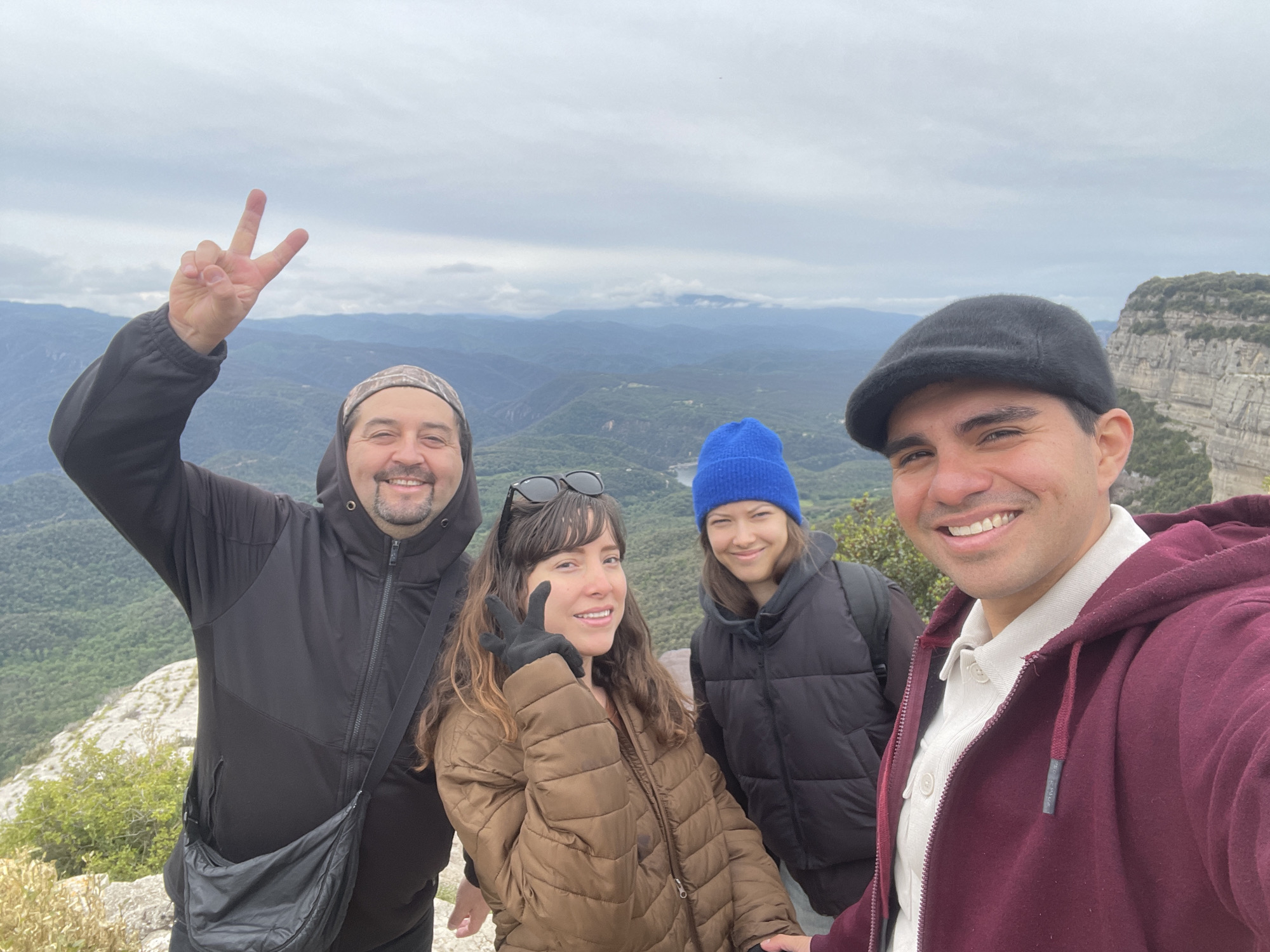 Four people are taking a selfie on a cliff in Tavertet, Spain. The man on the left is wearing a black hooded jacket, patterned beanie, and a crossbody bag, and is holding up a peace sign. Next to him, a woman is wearing a brown puffer jacket and sunglasses on her head, also holding up a peace sign. Beside her, another woman is wearing a blue beanie and a black puffer vest. On the right, the man taking the photo is wearing a black cap and a maroon hooded jacket. They are all smiling. The background features a mountain range covered in greenery, with a cloudy sky above. A rocky cliff is visible to the right.