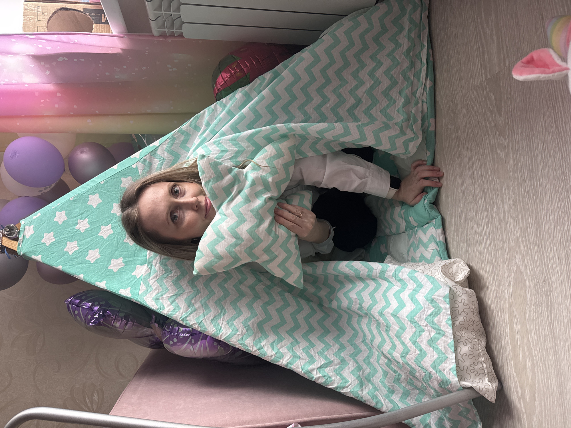 This image captures a young woman playfully emerging from a blanket fort within a cozy indoor setting in Chaikovsky, Russia. She has light hair and fair skin, looking directly at the camera with a warm, gentle smile. She is dressed in a white long-sleeved shirt, wearing a ring on her left ring finger and a smartwatch on her left wrist.
The fort is constructed from two main blankets: the one covering her is light mint green with white chevron stripes, while the "ceiling" of the fort is light mint green with white star patterns. She holds a small, matching chevron-patterned pillow.
The scene is festive, with various balloons decorating the space. Inside the fort, a red patterned foil balloon is partially visible behind her, and several round, solid-colored balloons in purple, grey, and white are seen in the background. Outside the fort, a large purple foil balloon with white patterns is also visible.
The surrounding environment includes a light-colored wooden floor to the right, a section of pink/purple upholstered furniture beneath the fort, and a white radiator on the wall. Wallpaper with a subtle swirling design is visible on the left. A plush toy, possibly a unicorn with pink and rainbow ears, is partially seen on the wooden floor to the right. Faint text "333-252-012" is visible on a cardboard box in the top left corner. The image conveys a joyful and comfortable atmosphere, suggesting a playful moment or celebration during the daytime.