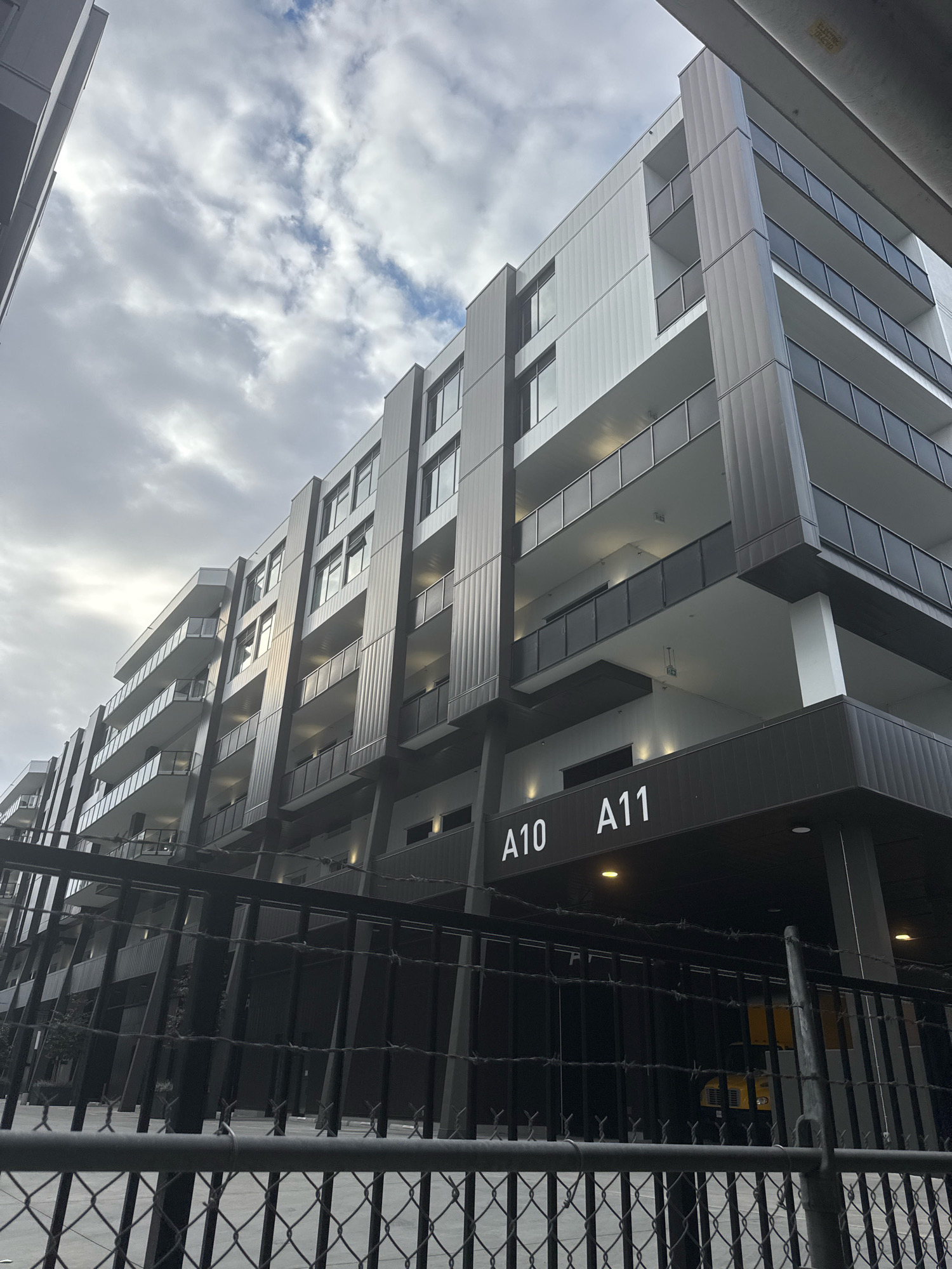 The image shows a modern, multi-story apartment building in Vancouver, Canada. The building features a facade of white and dark gray panels, with multiple balconies extending outwards. The sky above is overcast with clouds. In the foreground, a black metal fence with barbed wire runs across the bottom of the frame, partially obscuring the building. White lettering on a dark sign on the building reads "A10 A11". The scene appears to be an outdoor urban environment.