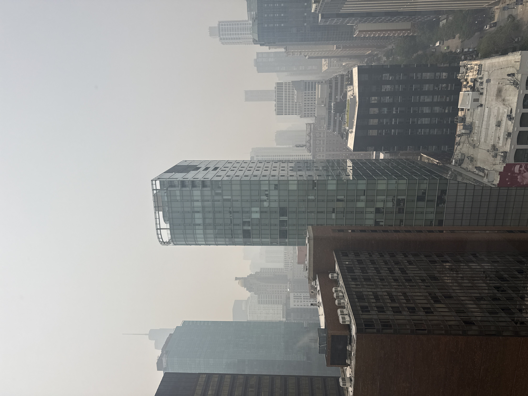 The image shows a hazy, overcast day in Chicago, United States of America, with numerous skyscrapers visible. The sky is a uniform light gray, obscuring the upper portions of many buildings and suggesting poor air quality, possibly due to smoke. The buildings are of varying heights and architectural styles, with a mix of modern glass towers and older brick structures. In the foreground, a dark brown brick building with many windows takes up the left side of the frame. To its right is a tall, slender glass skyscraper. Further back and to the right, another dark-colored modern building is prominent. The scene is dominated by the dense urban landscape and the pervasive haze. No people or specific actions are discernible.