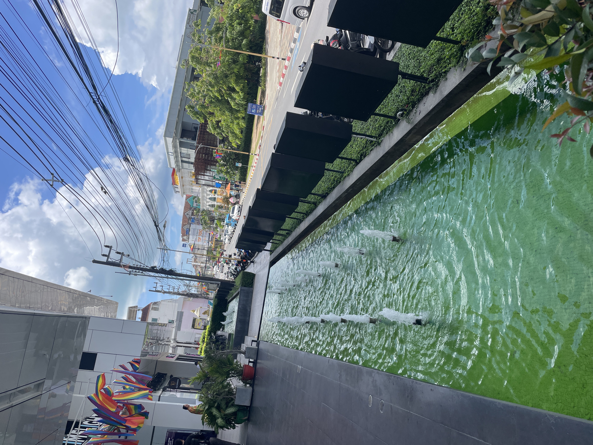 The image shows a street scene in Phuket City Municipality, Thailand, on a sunny day with scattered clouds. In the foreground, a long, narrow water feature with multiple fountains flows along the edge of a modern building. The water is a vibrant green color, and the bottom is tiled. To the left of the water feature is a dark gray tiled walkway. Adjacent to the walkway and the water feature is a strip of green bushes.
Beyond the water feature and bushes, a street runs parallel to the building. Cars and motorcycles are visible on the street, suggesting traffic. On the far side of the street are various buildings of different architectural styles and colors, including some multi-story structures with visible signage. Trees and streetlights are also present along the street. Overhead, a dense network of electrical wires crisscrosses the sky, supported by utility poles.
In the mid-ground, on the left side of the image, a person in a patterned shirt is standing near some potted plants. Flags, including rainbow flags, are displayed on the building. The overall impression is a busy urban environment with a mix of modern and traditional elements.