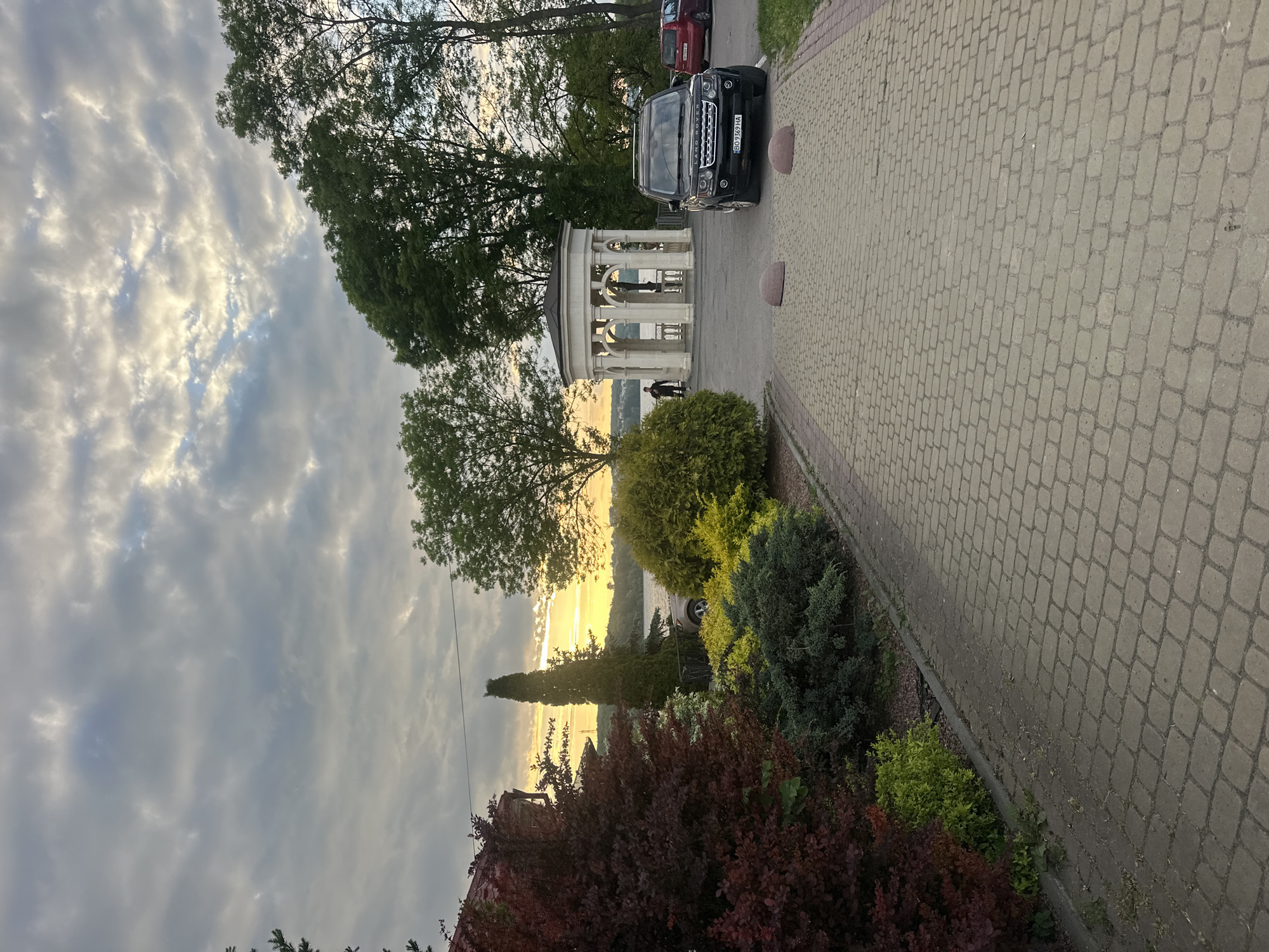 A scenic view in Ternopil, Ukraine, shows a paved walkway with a black SUV parked beside it. Beyond the walkway, ornamental shrubs and trees fill the landscape. A gazebo is visible, with two individuals standing nearby. A body of water reflects the setting sun in the background. The sky is filled with clouds.