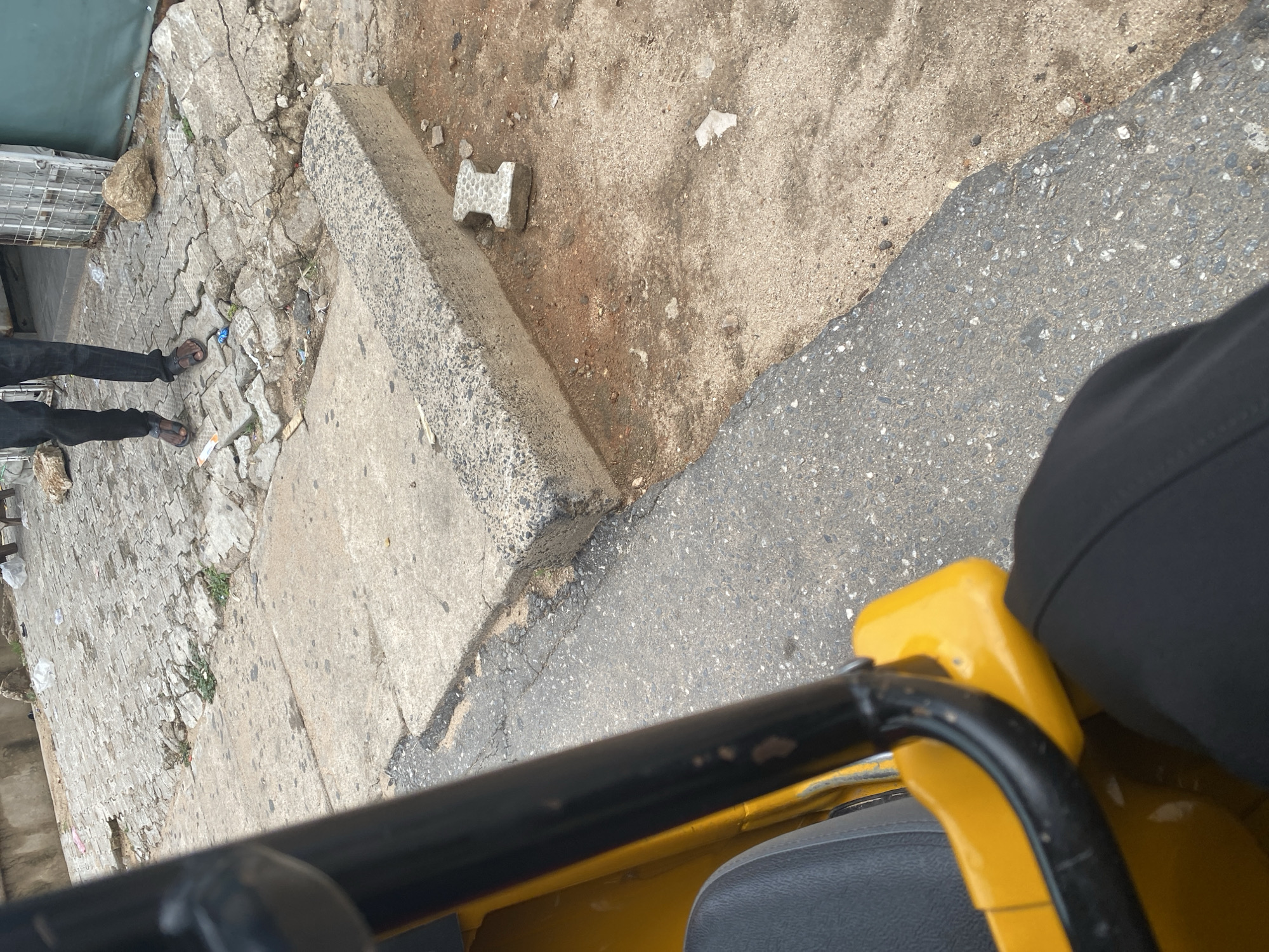 The image shows a low-angle, first-person perspective from a yellow vehicle, likely a tricycle or auto-rickshaw, looking forward and slightly to the left. The foreground is dominated by the yellow body of the vehicle and a black handlebar.
The scene is outdoors on a paved surface that transitions from rough concrete blocks and dirt to asphalt. To the left, a person's legs are visible, wearing black pants and dark sandals. The person appears to be standing or walking on the paved area. Further left, there are glimpses of a metal cage-like structure and what looks like a green tarp. Scattered debris and small rocks are present on the ground.
The ground itself shows signs of wear and tear, with cracked paving stones and patches of dirt. A distinctive H-shaped concrete block lies on the ground in the middle distance. The lighting suggests it is daytime, with no strong shadows indicating a specific time of day. The overall impression is of an urban or semi-urban environment in Jalingo, Nigeria, possibly a street or market area. There is no visible text in the image.