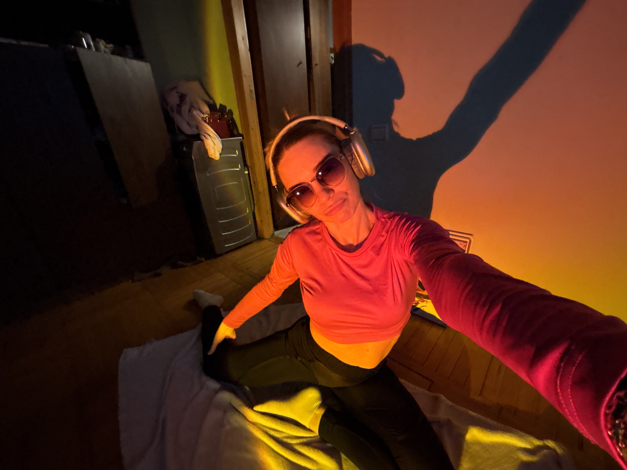 A woman is taking a selfie in what appears to be an indoor setting, possibly a bedroom or living space. She is sitting on the floor, which is partially covered by a light-colored blanket or rug. She is wearing a pink long-sleeved crop top, dark leggings, sunglasses, and headphones. She is in a split position, with one leg extended forward and the other backward. The woman is angled slightly to the left of the frame and is holding the camera or phone in her right hand, extending her arm towards the viewer.
The lighting in the room appears to be a mix of natural and artificial, with a warm, golden hue. The wall behind her is colored in shades of orange and yellow, and there is a dark shadow that resembles a person standing with their arm raised. In the background, there are pieces of furniture, including what seems to be a chest of drawers with some items on top. The overall composition suggests a casual and personal moment captured indoors. The location of the city and the details are unknown.