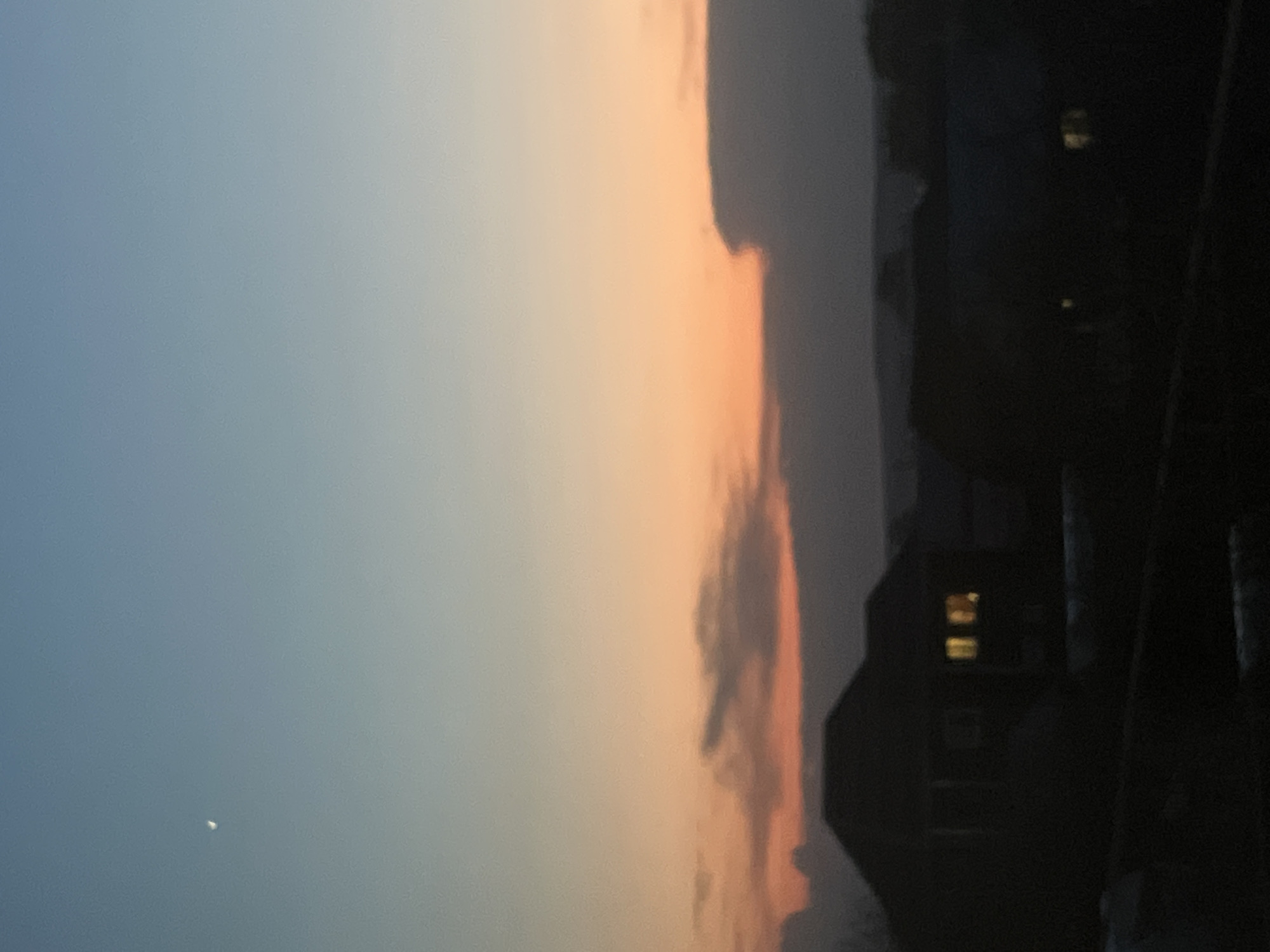 The image captures a dusk or dawn scene. On the left, the sky transitions from blue to a warm orange hue. In the lower left corner is a small, bright celestial body, possibly the moon or a star. Toward the right, dark, silhouetted buildings are visible, one with illuminated windows. The exact location in Votkinsk, Russia, cannot be determined from the visual information provided. There are no people, subjects, actions, or interactions visible in the image. The environment appears to be urban or suburban.
