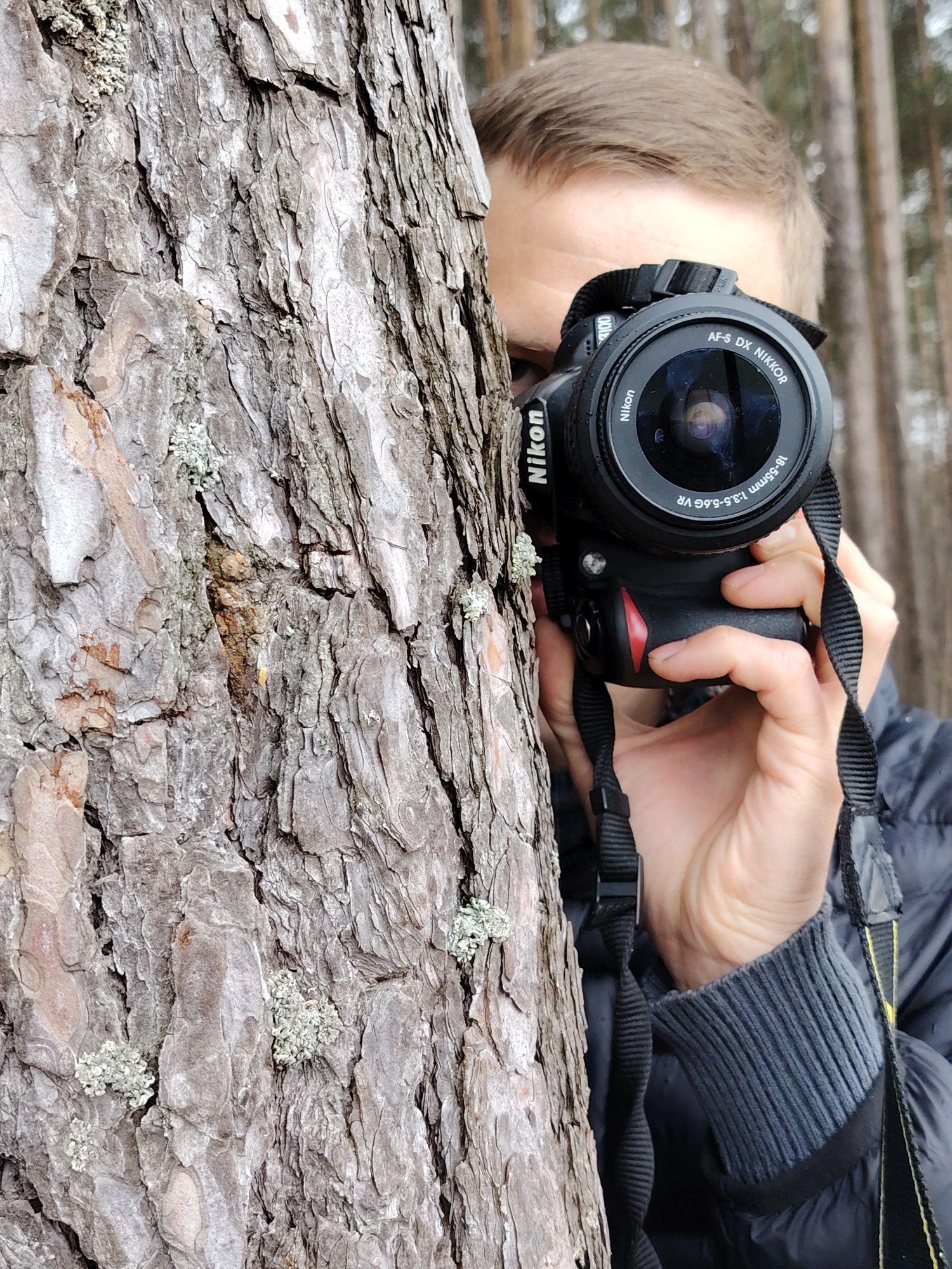 A man is half hidden behind a tree while holding a Nikon camera. The camera is black with a Nikon lens, and the lens reads "AF-S DX NIKKOR 18-55mm 1:3.5-5.6G VR." The man is wearing a dark-colored jacket with a grey ribbed sleeve visible. The tree has light grey bark. The background is blurred, but appears to be a forest setting with trees and a neutral-colored ground. Based on the location of Chaikovsky, Russia, the environment is likely a natural, forested area. There is no other information to specify the exact location.