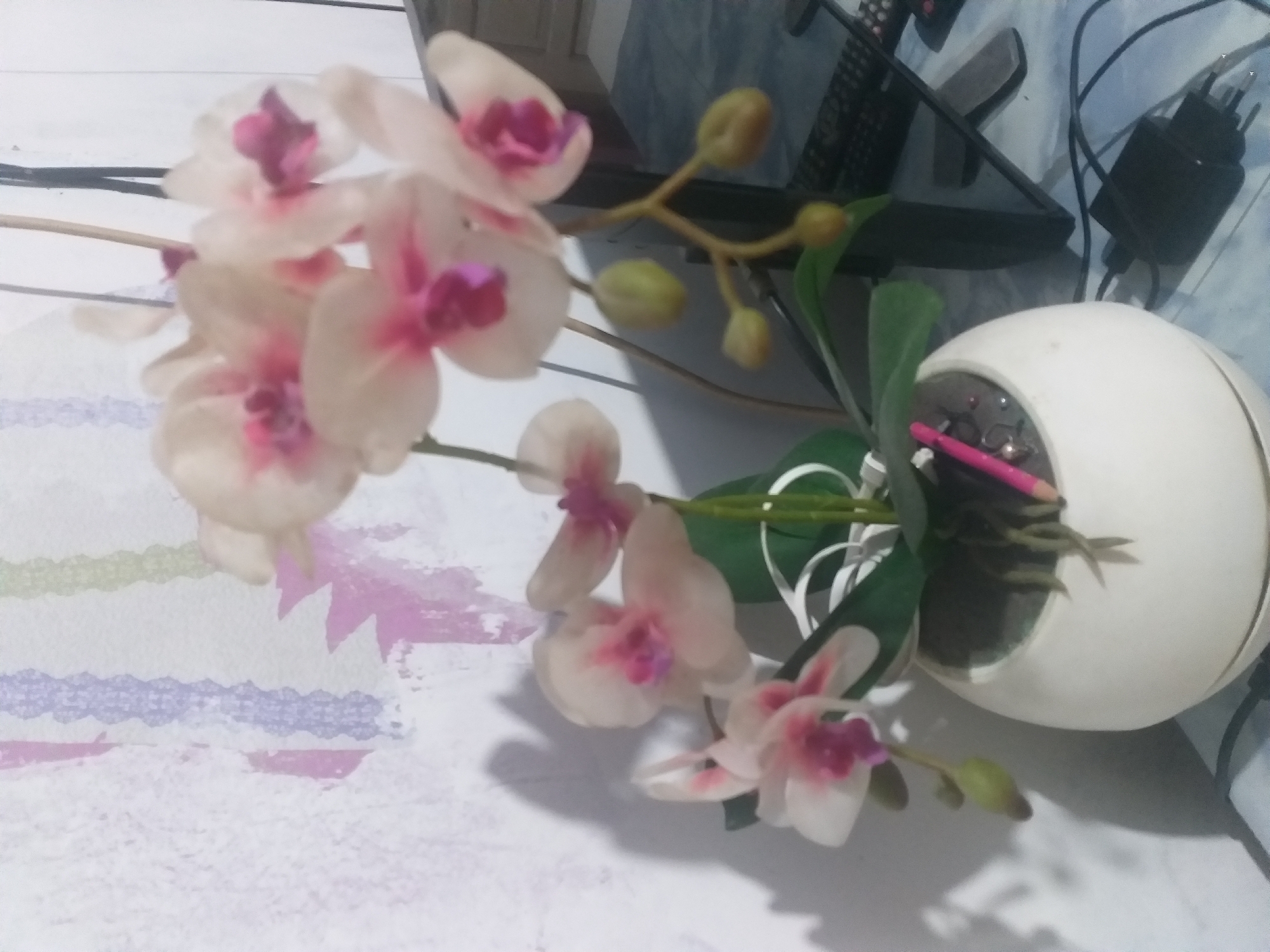 The image shows a potted artificial orchid plant with white and pink flowers. The plant is in a white, round pot, and there are green leaves and several yellow buds. A pink pencil is stuck in the soil of the pot. The background consists of a wall with a striped pattern and a shelf with electronic devices, including a keyboard and a power adapter. The scene appears to be indoors, possibly in a home or office. There is no information available to determine the location in the city of Dumai City, Indonesia.
