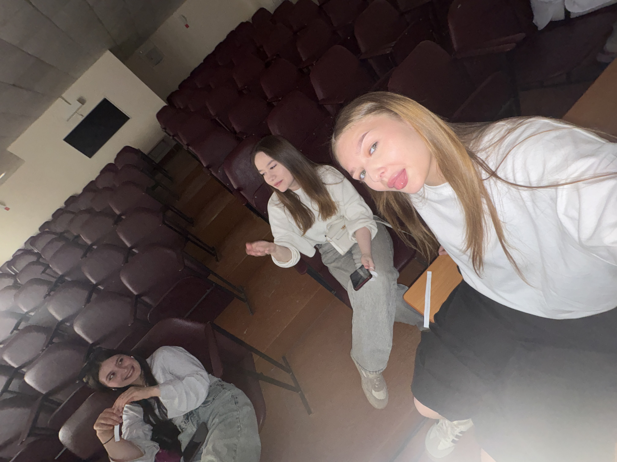 Three young women are in a large auditorium with rows of maroon seats. The auditorium is likely located in Chaikovsky, Russia. The woman on the right is taking a selfie and sticking out her tongue. She is wearing a white long-sleeved shirt and a black skirt. The woman in the middle is wearing a white top, gray pants, and white sneakers, and is holding a cell phone in her hand. The woman on the left is lying on the seats, smiling. She is wearing a white top and jeans. The lighting in the auditorium is dim.