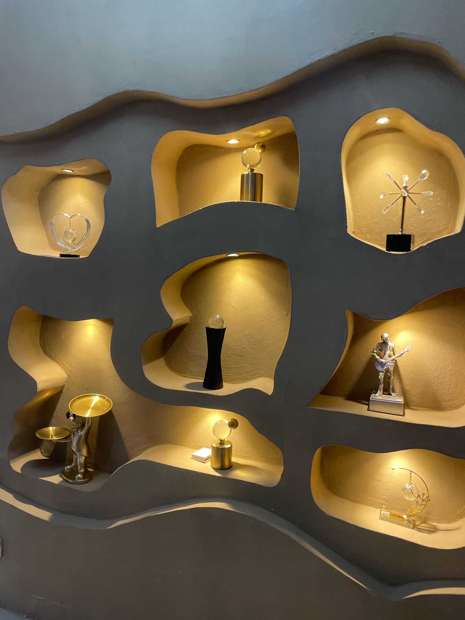 The image shows a wall with recessed niches decorated with various objects. The wall has a textured, organic, wavy design. The niches are illuminated by spotlights, casting a warm glow on the displays.
In the top left niche, there is a transparent heart-shaped sculpture with intricate swirling details, resting on a black base.
To the right of the heart sculpture, in the next niche, a cylindrical gold base supports a clear spherical object, possibly a crystal ball, with a smaller sphere on top.
The top right niche features a metallic sculpture resembling a starburst or atom, with radiating rods tipped with small crystal-like elements, mounted on a black base.
Below the heart sculpture, in the middle left niche, a dark, elongated vase-like object with a textured surface stands prominently.
In the middle niche, a clear sphere with a gold cap sits atop a cylindrical gold base. To its left, a small white rectangular object is visible.
In the middle right niche, a silver-toned figurine of a person playing a guitar is displayed on a silver rectangular base.
In the bottom left niche, a metallic figurine of a person holding a tray and a smaller dish is positioned. The figurine appears to be wearing sunglasses.
The bottom right niche contains a delicate gold metal sculpture with a crescent moon shape, adorned with clear crystalline elements and a small gold leaf motif.
The overall color palette is dominated by neutral grays and warm golds, with the metallic and crystalline objects providing highlights. The lighting creates a dramatic effect, emphasizing the textures and forms within the niches. This scene is located in the city of Ibadan, Nigeria.