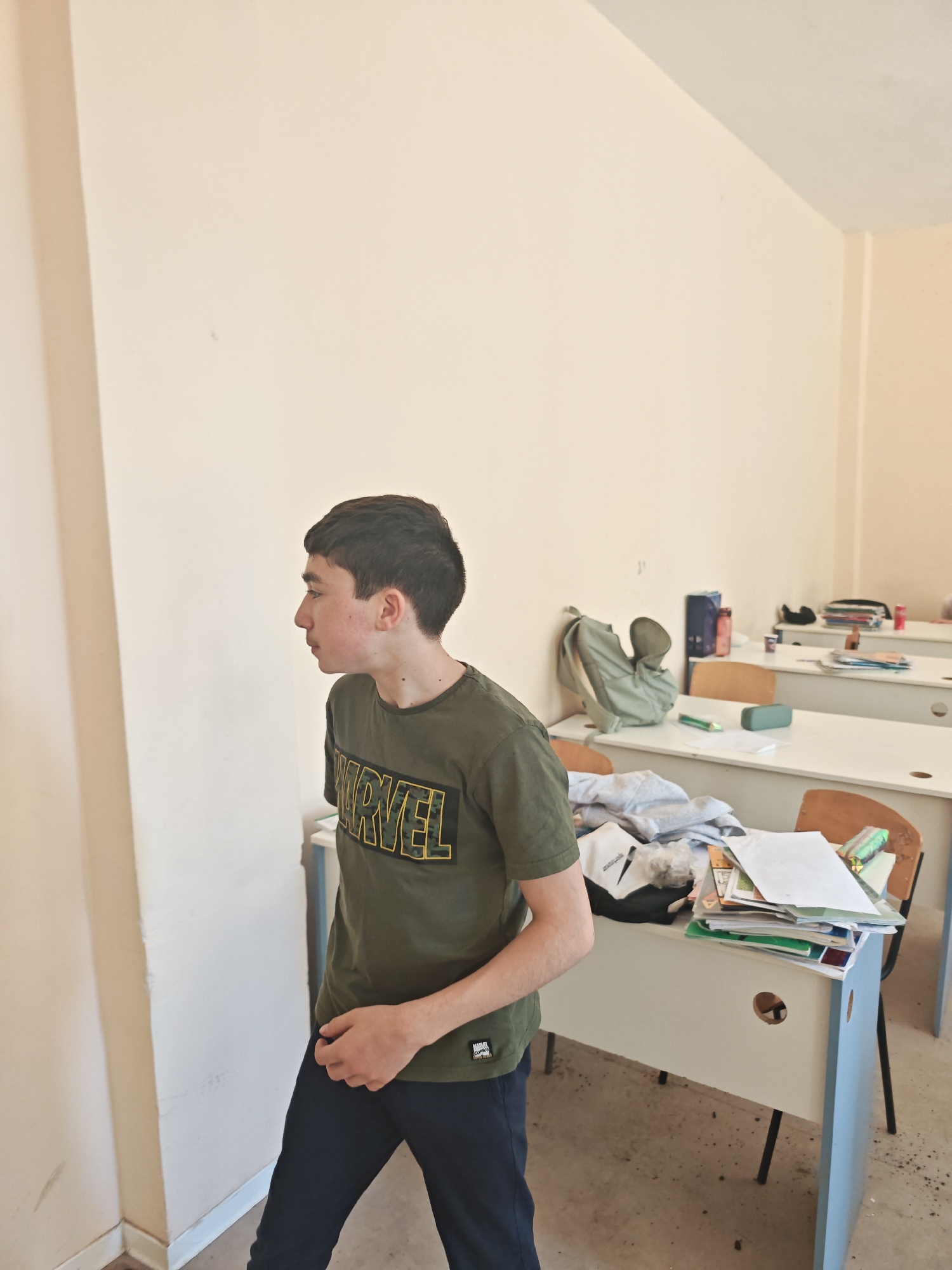 A young male, likely a teenager, stands in profile, looking towards a plain light-colored wall in what appears to be a classroom in Varna, Bulgaria. He has short dark hair and wears a dark olive green t-shirt with "MARVEL" printed in yellow, camouflaged letters across the chest, and dark trousers. His expression is neutral, suggesting contemplation or observation.
The background reveals a classroom environment with several light-colored desks, featuring white tops and blue side panels, paired with wooden chairs. The desks are somewhat cluttered with various items: a green backpack, notebooks, loose papers, a green pencil case, and a red water bottle. One desk also holds what appears to be folded clothes or cleaning materials. The walls are a creamy beige, and the floor is a light-colored, possibly concrete, surface with some marks. The room is well-lit, indicating it is daytime. A small, illegible text label is visible on the bottom left hem of his t-shirt. The overall impression is a candid shot within an educational setting.
