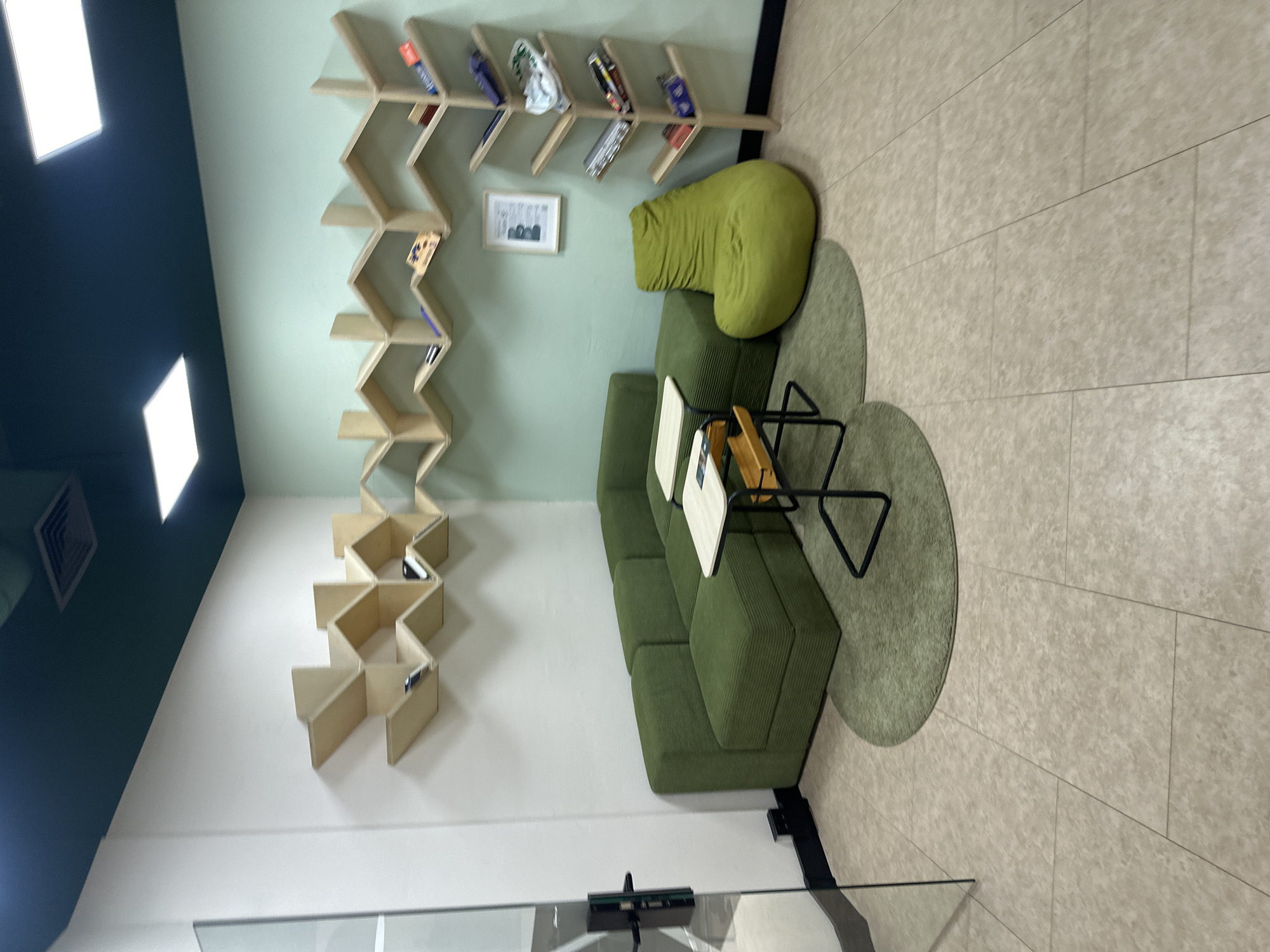 The image depicts an interior space, possibly a common area or lounge, in Budva, Montenegro. The room features a light green wall with a zig-zag wooden bookshelf containing books and objects. A green corduroy sofa and a green beanbag chair are positioned near the bookshelf. A small table with a black frame sits on a round green rug. The floor is beige tile. The lighting is bright, with recessed ceiling lights visible in the dark-colored ceiling. There are no people visible in the frame. A framed picture hangs on the wall between the bookshelf and the beanbag chair. A glass barrier is at the bottom of the frame.