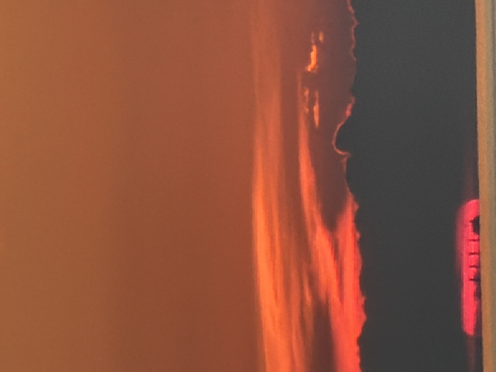 This is a photograph of a sunset over a body of water. The sky is filled with vibrant orange and red hues, with layers of clouds creating depth and texture. The sun, a bright red disc, is partially obscured by a dark silhouette of clouds at the horizon. In the foreground, a thin sliver of water is visible, reflecting the colors of the sky. Silhouetted against the sun and water are several indistinct vertical shapes, which appear to be ships or structures. The overall impression is one of a dramatic and serene natural spectacle.