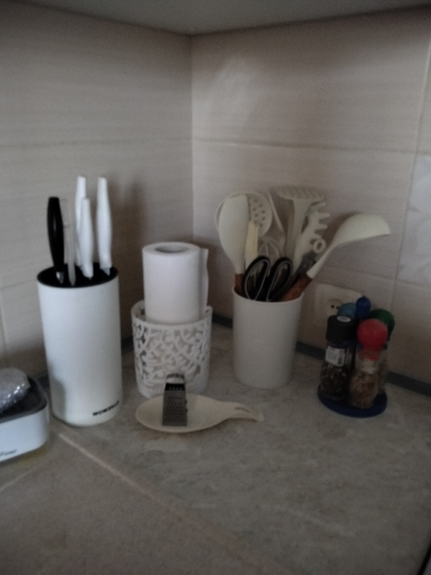 In a kitchen environment, a white cylindrical knife block holds several knives, one with a black handle and the others with white handles. To the right of the knife block is a roll of white toilet paper placed in a white decorative holder. Further to the right, a white cup contains various kitchen utensils, including what appears to be a whisk, spatulas, and scissors with black handles. Adjacent to the cup is a set of salt and pepper shakers, with a blue base. In the foreground, a small white grater rests on a light-colored dish. The kitchen counter is a neutral gray color, and the backsplash is tiled in a light beige. The overall scene depicts organized kitchenware.