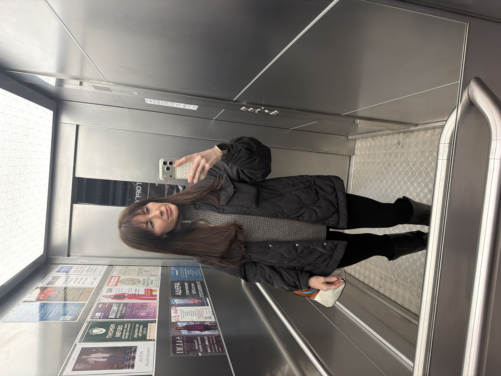 The photograph shows a woman taking a selfie in what appears to be an elevator. She is holding a smartphone in her right hand, facing a mirror. She is wearing a black quilted jacket, a gray sweater, black pants, and black boots. In the elevator, there are advertisements posted on the wall to the left. Based on the text in the advertisements, the location of the elevator is in Perm, Russia. The elevator has metal walls and a textured metal floor. A railing is visible on the right side of the elevator.
