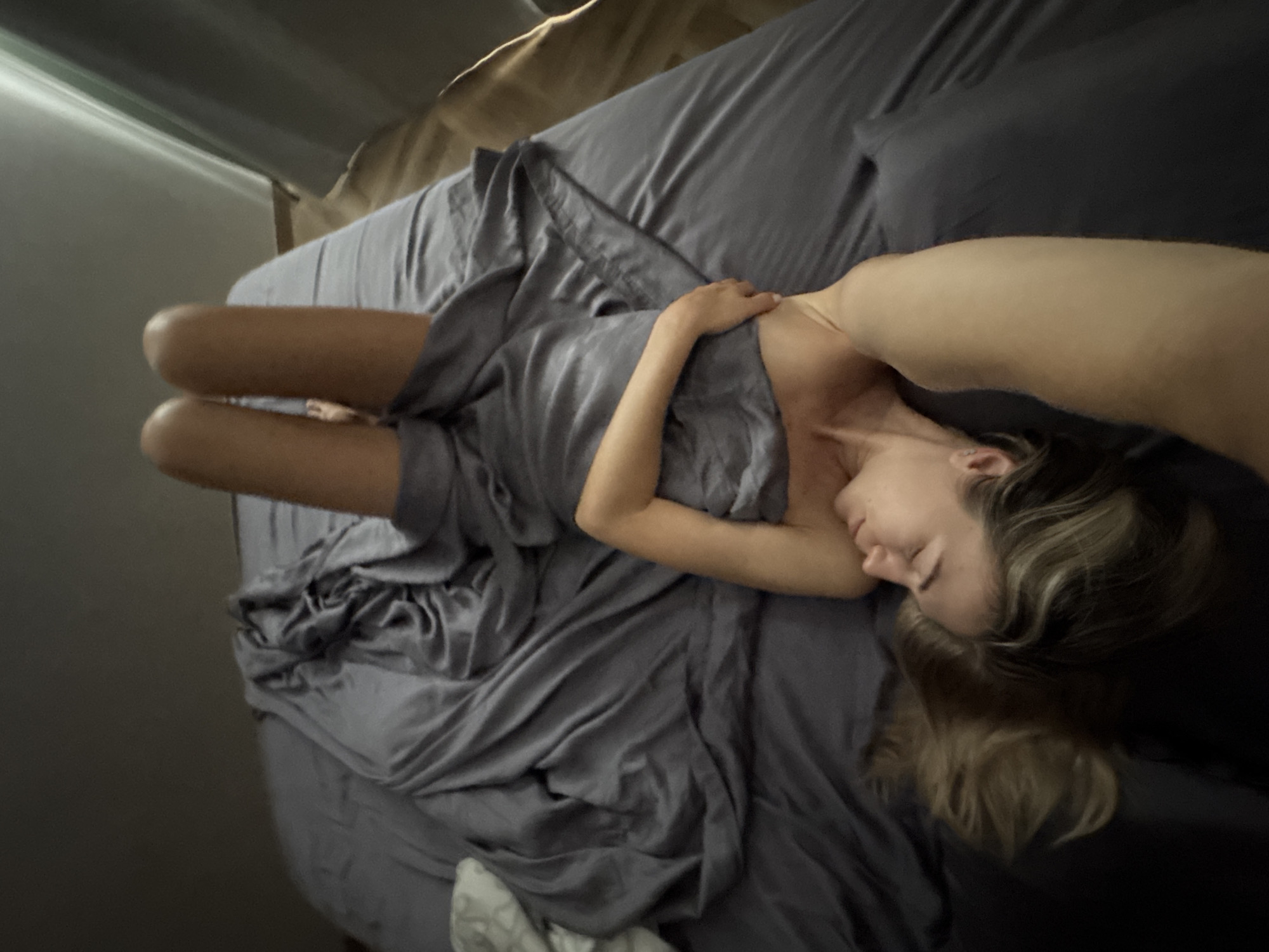 A woman is lying on a bed, covered by a grey sheet. Her legs are extended upwards, and her body is positioned on its back. Her head is tilted to the side, and her eyes are closed. Her hair is a mix of blonde and brown, and it is spread across the pillow. The bed is covered with a grey sheet, and there is a white pillow on the left side of the bed. The room is dimly lit, and the walls are a dark grey color. The image appears to be taken from a high angle, looking down at the woman on the bed.