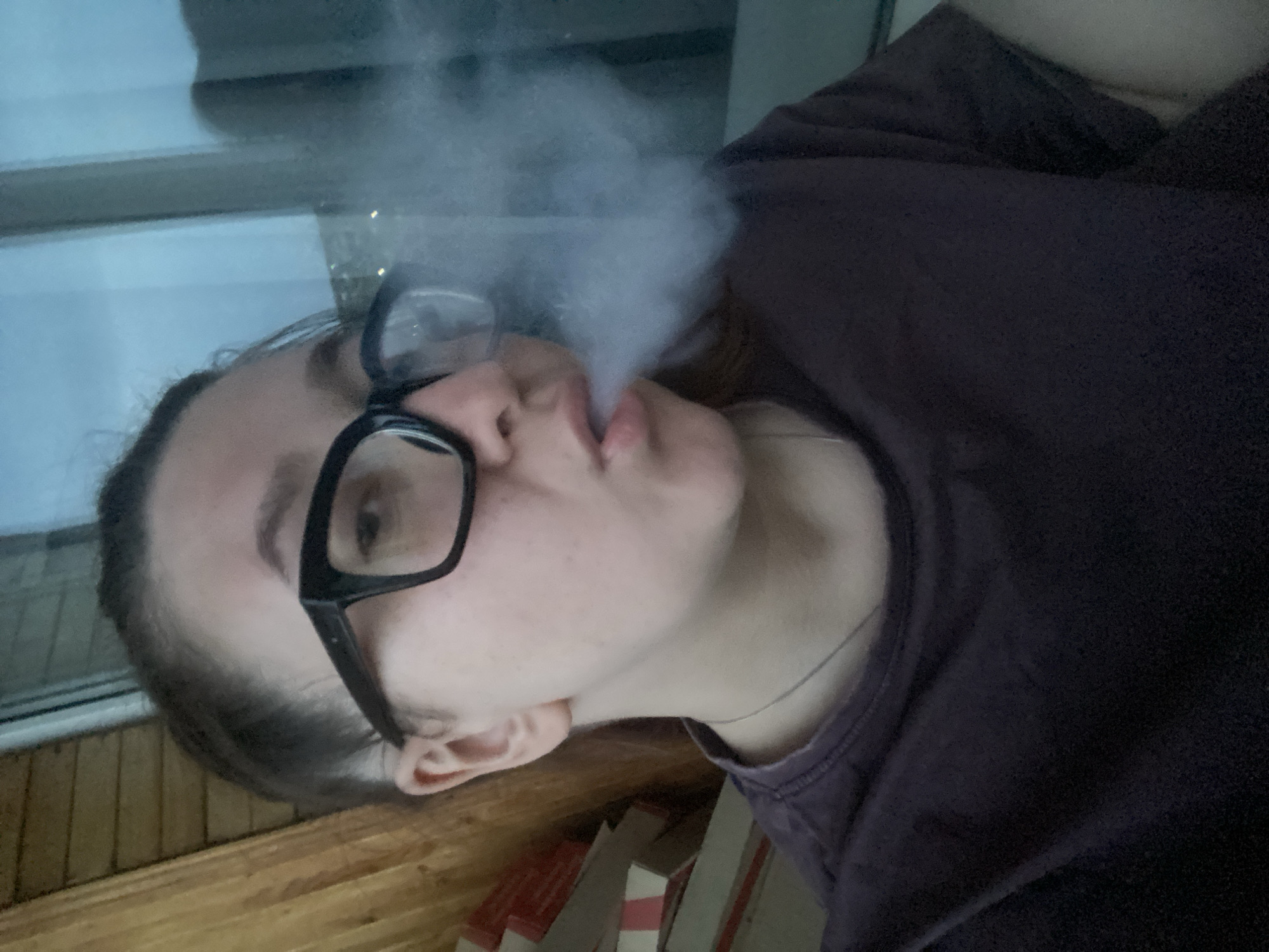 A person with glasses is exhaling smoke indoors. The person is wearing a dark-colored t-shirt and appears to be leaning against a wooden surface. A window is visible in the background. The location may be a balcony or an enclosed porch due to the appearance of a wooden railing or wall. The scene lacks specific identifying details to confirm the city of Chaikovsky.