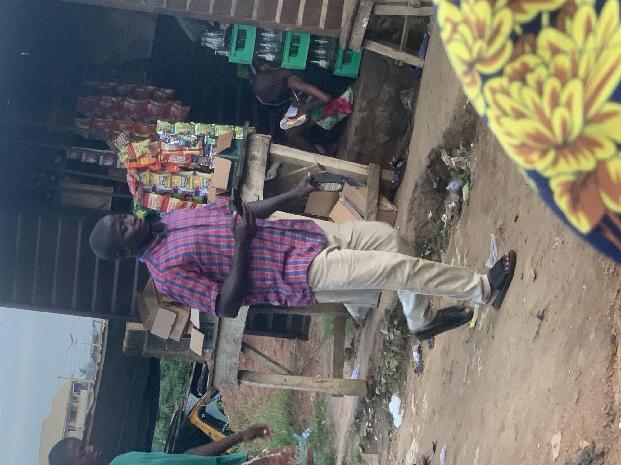The image depicts a scene in a dusty, outdoor market area in Shagamu, Nigeria. A man in a red and blue plaid shirt and light-colored pants is walking towards the viewer, holding a smartphone in his left hand and a black object, possibly a drink bottle or a remote, in his right hand. He is wearing black sandals. To the left, partially visible, is another person wearing a green shirt. Behind the man in plaid, a makeshift shop is set up with shelves displaying various packaged snacks and drinks. A person is seated inside the shop, appearing to be engaged with something on their lap. In the background, there is a building and some vegetation. The ground is dirt and strewn with debris, including plastic bags and wrappers.