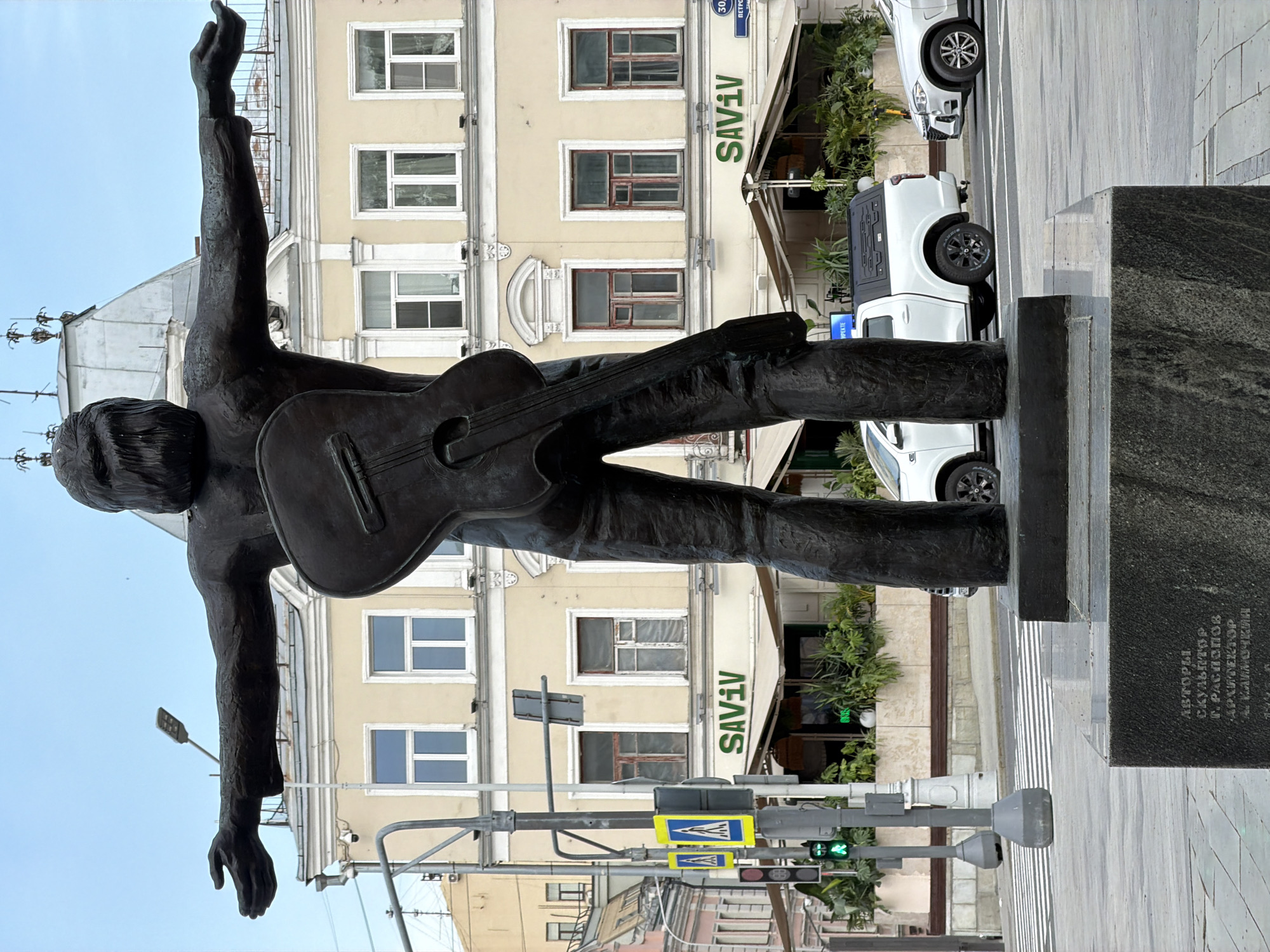 An upright-oriented image captures a bronze statue of a male musician holding a guitar and raising his left arm high, seemingly mid-performance, looking skyward. The statue stands on a dark, polished granite pedestal. Behind the monument, a light-colored, multi-story building with white window frames and decorative architectural elements lines an urban street. An awning on the ground floor of this building displays the text "SAVIV".
Several white cars, including a pickup truck, are parked along the street. The road surface features distinct white pedestrian crossing markings. Various street furniture is visible, including traffic lights (one showing green), a lamppost, and street signs. The sky is clear and light blue, indicating a bright daytime scene.
On the statue's pedestal, Cyrillic text is inscribed: "АВТОРЫ: СКУЛЬПТОРЫ Г. РАСПОПОВ АРХИТЕКТОР А. КАПУОКИН" (Authors: Sculptors G. Raspopov, Architect A. Kapuokin). A blue street sign in the background clearly shows "ПЕТРОВКА 30А" (Petrovka 30A), providing a specific location cue for this Moscow street. Yellow pedestrian crossing signs are also visible on poles.