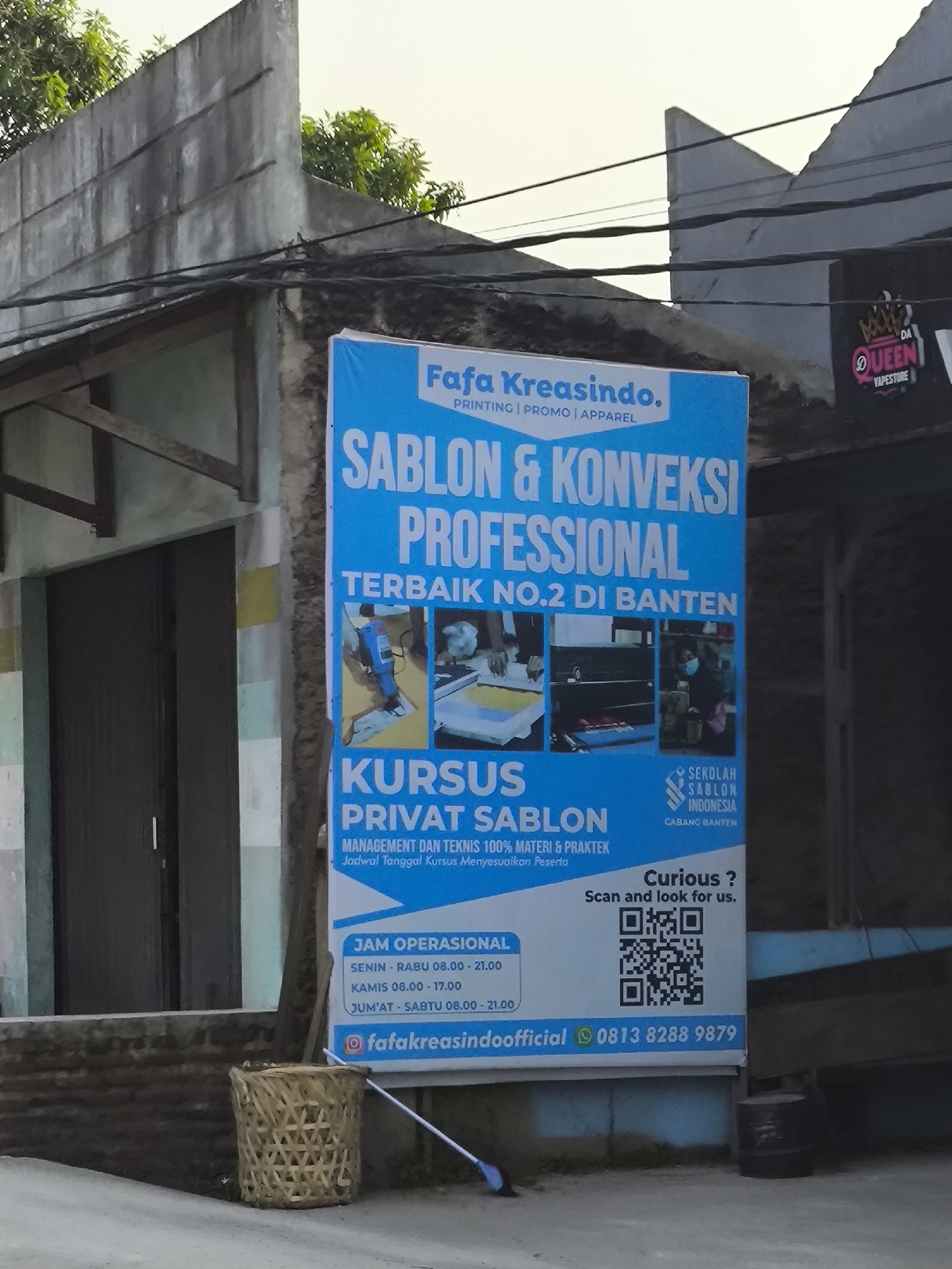 The image captures a vibrant blue banner advertising "Fafa Kreasindo" printing, promo, and apparel services, located in Cilegon, Indonesia. The banner highlights "Sablon & Konveksi Professional" and offers "Kursus Privat Sablon" (Private Screen Printing Courses). Below the course details, "Jam Operasional" (Operational Hours) are listed from Monday to Sunday. A QR code is prominently displayed with the text "Curious? Scan and look for us." and contact information, including an Instagram handle and a phone number. The banner is mounted on the exterior of a building, partially obscuring a dark-colored garage door. To the left of the banner, a woven bamboo basket sits on the ground, and a white broom lies nearby. In the background, overhead electrical wires crisscross against a bright sky, and glimpses of trees and other buildings are visible. To the right of the banner, another smaller sign with a "Queen Vapestore" logo is partially visible. The scene suggests an outdoor commercial setting in a moderately urban environment.