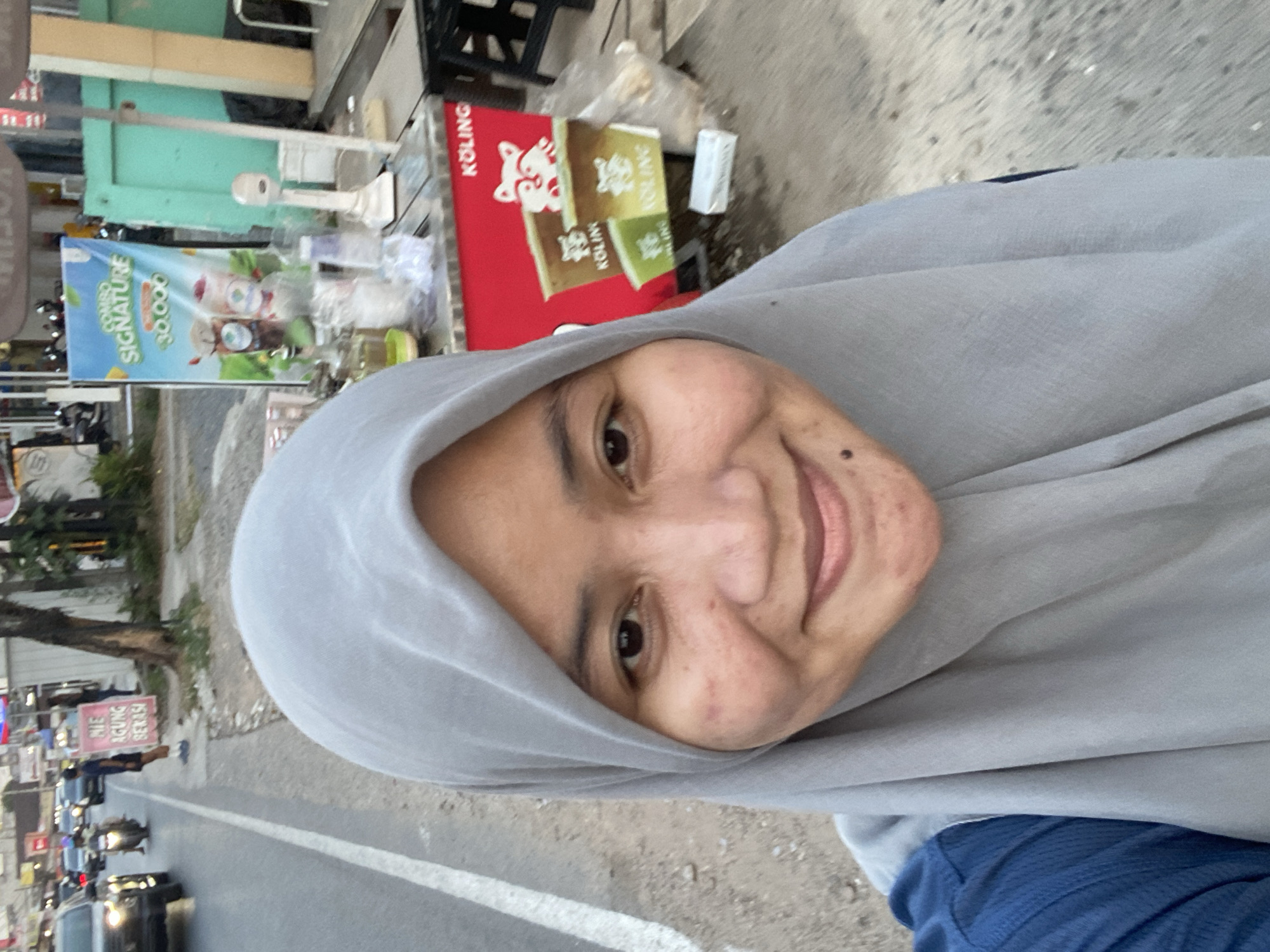A medium shot captures a woman wearing a light gray hijab and a blue shirt, smiling at the camera in what appears to be an outdoor market setting in Banjarbaru, Indonesia. The background features a red food cart with "KOLING" branding, displaying colorful cups with cartoon raccoons. To the left of the cart, a blue sign with white text reads "MIE AGUNG BEKASI." Further in the background, a street with passing cars and motorcycles suggests an urban environment. The woman's face is the primary focus, with visible skin texture and light makeup. The overall scene is casual and appears to be taken during the daytime.