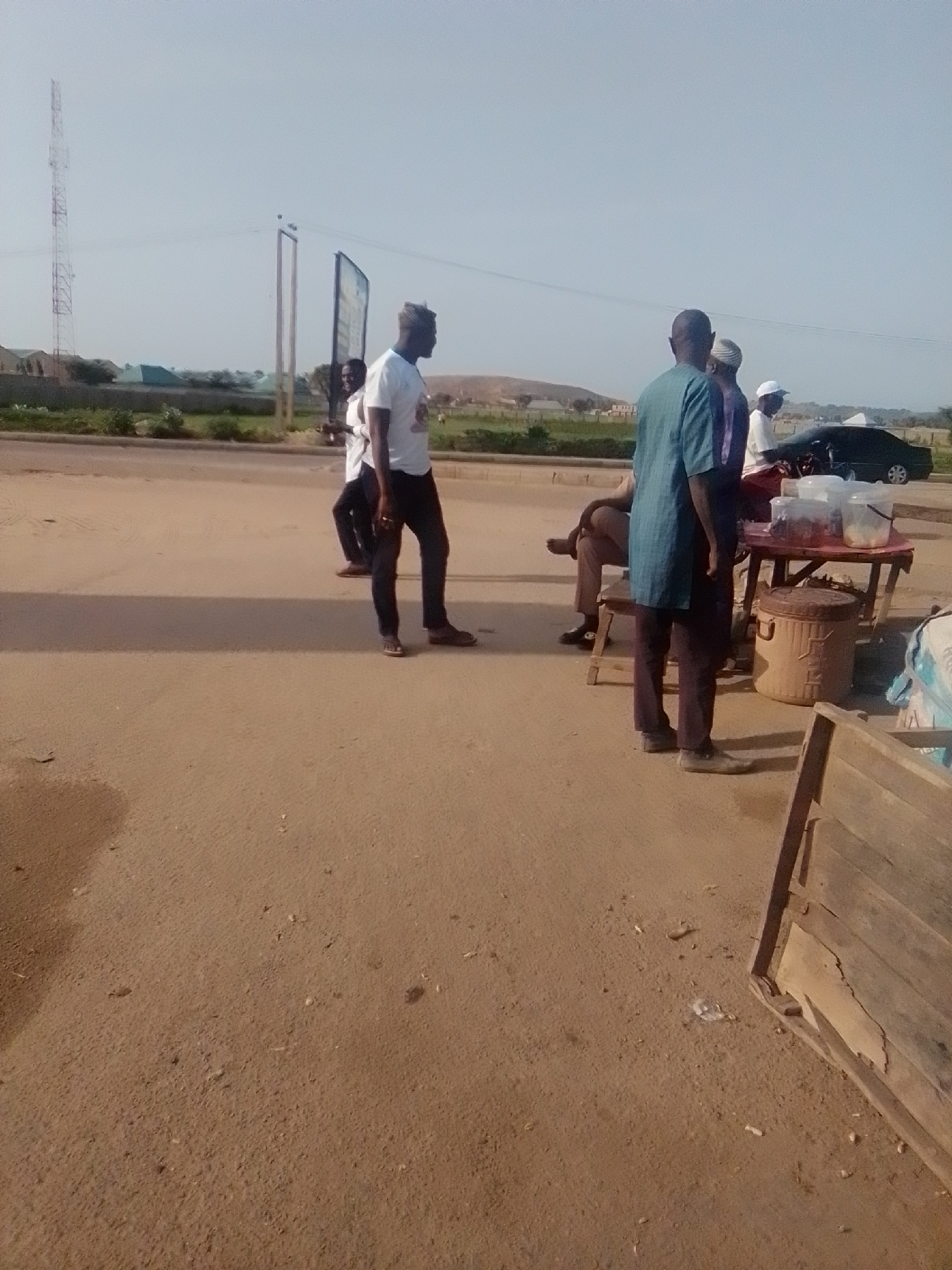 The image depicts a street scene in Gombe, Nigeria, under a clear, pale sky. Several individuals are present near a roadside stall laden with containers of food and drinks. A tall communication tower is visible in the distance to the left, alongside low-rise buildings and vegetation. The ground is a dusty, brown earth.
In the foreground, on the right, a makeshift wooden structure, possibly a table or stall, partially enters the frame. To its left, a round, metal trash bin with visible markings is situated. In the mid-ground, a red-topped table is set up, displaying an assortment of clear containers filled with food items. A black car is parked to the right of the table.
There are five men in the image, all appearing to be of African descent. One man, wearing a blue-green kaftan and dark trousers, stands facing away from the camera, looking towards the stall. Another man, in a purple and white traditional outfit, stands next to him. Further to the left, a man in a white t-shirt and black trousers is captured mid-stride, walking away from the camera. He appears to be the primary subject in the left half of the image. Another man in a white t-shirt is partially visible behind him. A fifth man, wearing a light-colored hat, is seated on a low stool near the table, facing the stall.
The overall atmosphere suggests a casual, outdoor gathering, likely a place where food and refreshments are sold. The presence of the communication tower and the style of dress indicate an urban or semi-urban setting within Nigeria.