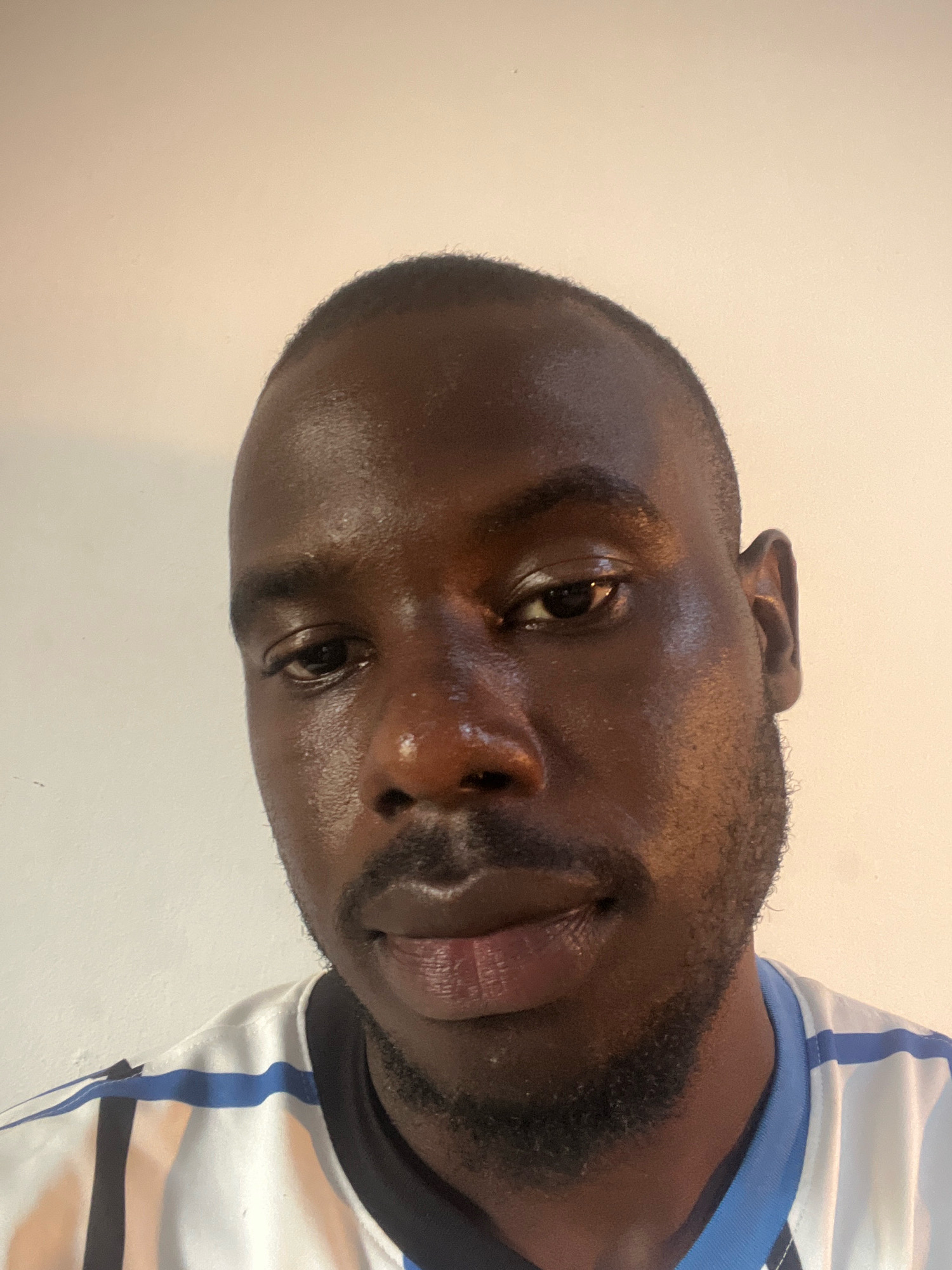 The image is a close-up, chest-up photograph of a Black man against a plain, light-colored wall. The man's head is tilted slightly downwards and to his left. He has dark skin, short-cropped hair, and a faint beard and mustache. His eyes are open and looking forward. He is wearing a white t-shirt with blue and black stripes across the chest and shoulders. The lighting is warm and casts subtle shadows on his face, highlighting the contours of his features. There is no discernible environment beyond the wall and the man's clothing. The image does not contain any information that would allow for the determination of its location in Gura Top, Nigeria, or any other specific geographical location.