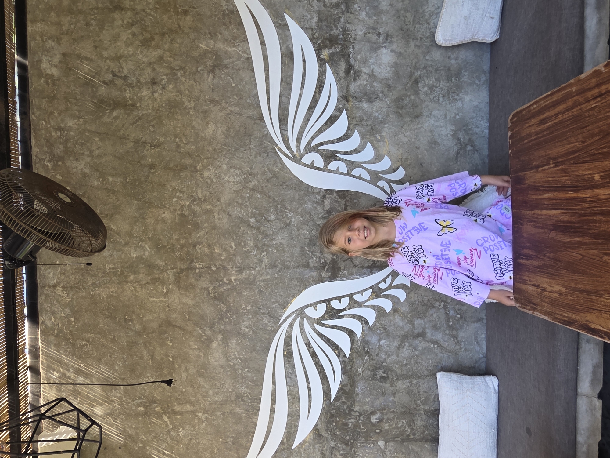 A girl with blonde hair is posing for a picture in front of a wall with a painted set of angel wings. The location is North Kuta, Indonesia. The girl is wearing a light purple, long-sleeved top with various graphic designs, including the words "Grow Positive." Her arms are partially outstretched to coincide with the angel wings. She is standing in front of a gray concrete wall with a large set of white angel wings painted on it. Next to her is a wooden table and what appears to be a white cushion on the ground. A ceiling fan and other architectural elements are visible in the background.