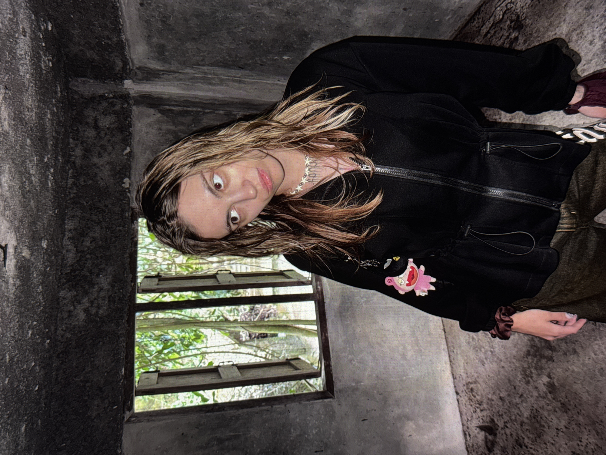 A young woman with wet, blonde hair stands in a dark, concrete room with a small window to her left. She is wearing a black zip-up jacket with a pink charm attached to the zipper. Her eyes are looking to the side, and she has a necklace on. The window shows lush green trees and vegetation. The overall scene suggests an indoor, possibly abandoned or neglected, location. There is no discernible information about the city of Moscow, Russia, or any specific actions or interactions occurring.