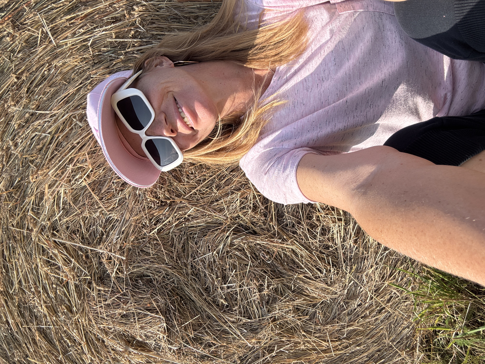 A woman with blonde hair, wearing a pink baseball cap and white-framed sunglasses, sits outdoors leaning against a large round hay bale. She is wearing a light pink t-shirt and black pants. The background consists of the rough texture of the hay bale, with some green grass visible at the bottom left. The lighting suggests it is daytime, with sunlight casting shadows on her face and t-shirt. The image is a close-up selfie. The location is Yablonovka, Russia.