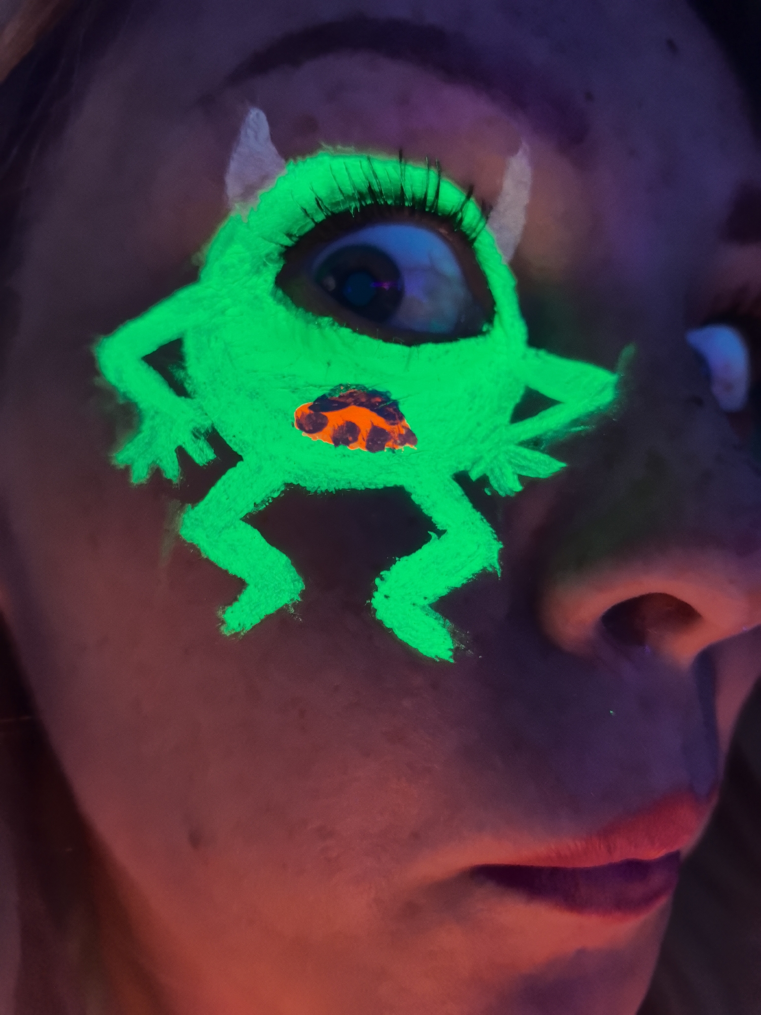 A close-up image shows a person's face illuminated by UV light, highlighting vibrant, fluorescent makeup. A cartoon-like creature, resembling a green monster, is painted across the person's right eye and cheek. The person's right eye serves as the monster's large, central eye, complete with visible black eyelashes and a blue iris. Small, white, horn-like shapes are painted above the monster's eye, extending onto the forehead. The monster features a body with four limbs and an expressive mouth area in fluorescent orange with black accents, glowing brightly against the dark background.
The person's skin not covered by the glowing paint appears in reddish-purple tones under the UV illumination. On the right side of the frame, parts of the person's nose and lips are visible; the lips are painted with a reddish lipstick that also exhibits a subtle glow. The background is indistinct and dark, emphasizing the glowing makeup. The image captures a creative makeup application, likely for an event such as a party or artistic performance under blacklight conditions. No text is visible. This scene takes place in Noviy, Russia.