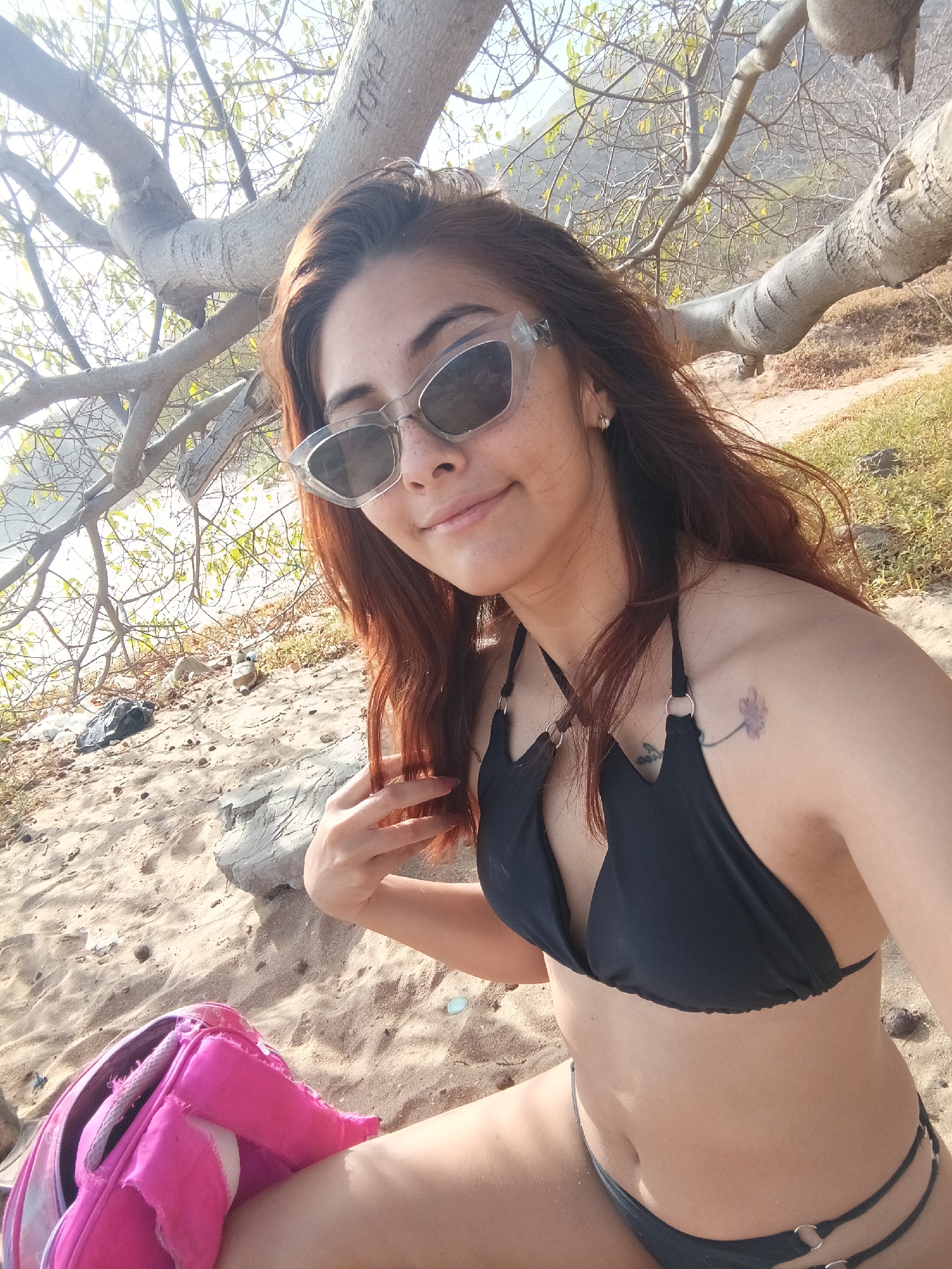 The photograph depicts a woman in a black bikini, seated on a sandy beach under the partial shade of a tree. She is wearing sunglasses and has reddish-brown hair. A pink backpack is visible near her on the sand. In the background, there's a glimpse of water and vegetation. The image was taken in Pedro González, Venezuela.