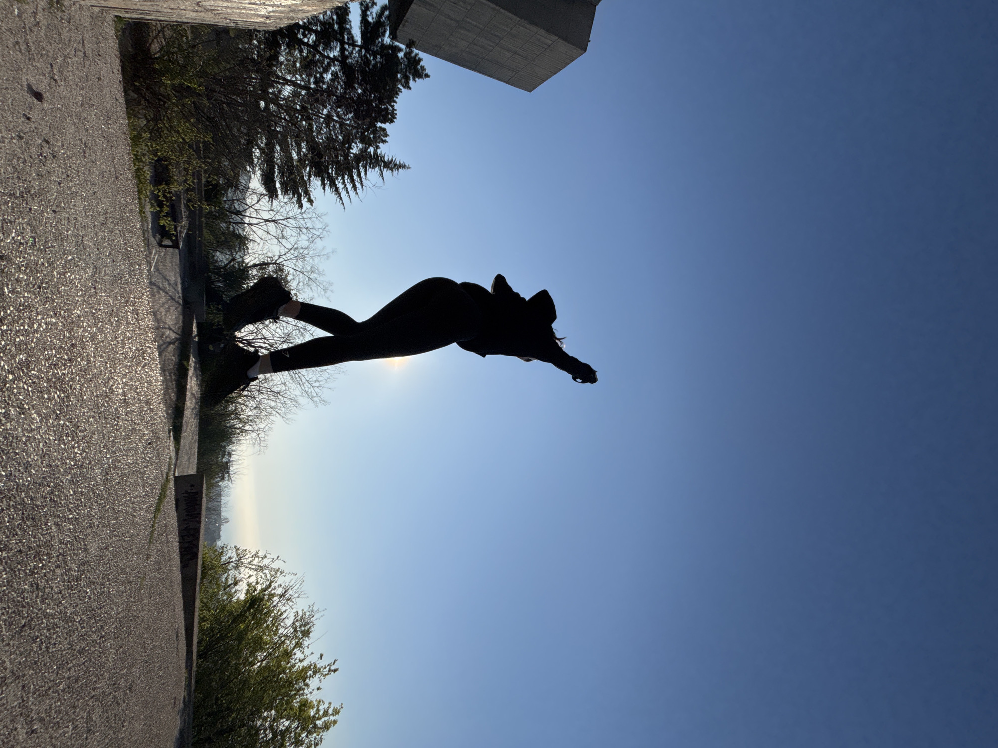 A silhouette of a person with outstretched arms against a blue sky, potentially performing a dive or jump. They are wearing dark clothing. The person is positioned near a concrete structure, potentially a building or monument. There are trees and greenery visible along the edge of the frame, suggesting the setting is outdoors, possibly a park or urban area. The location is specified as "Unknown, Unknown".