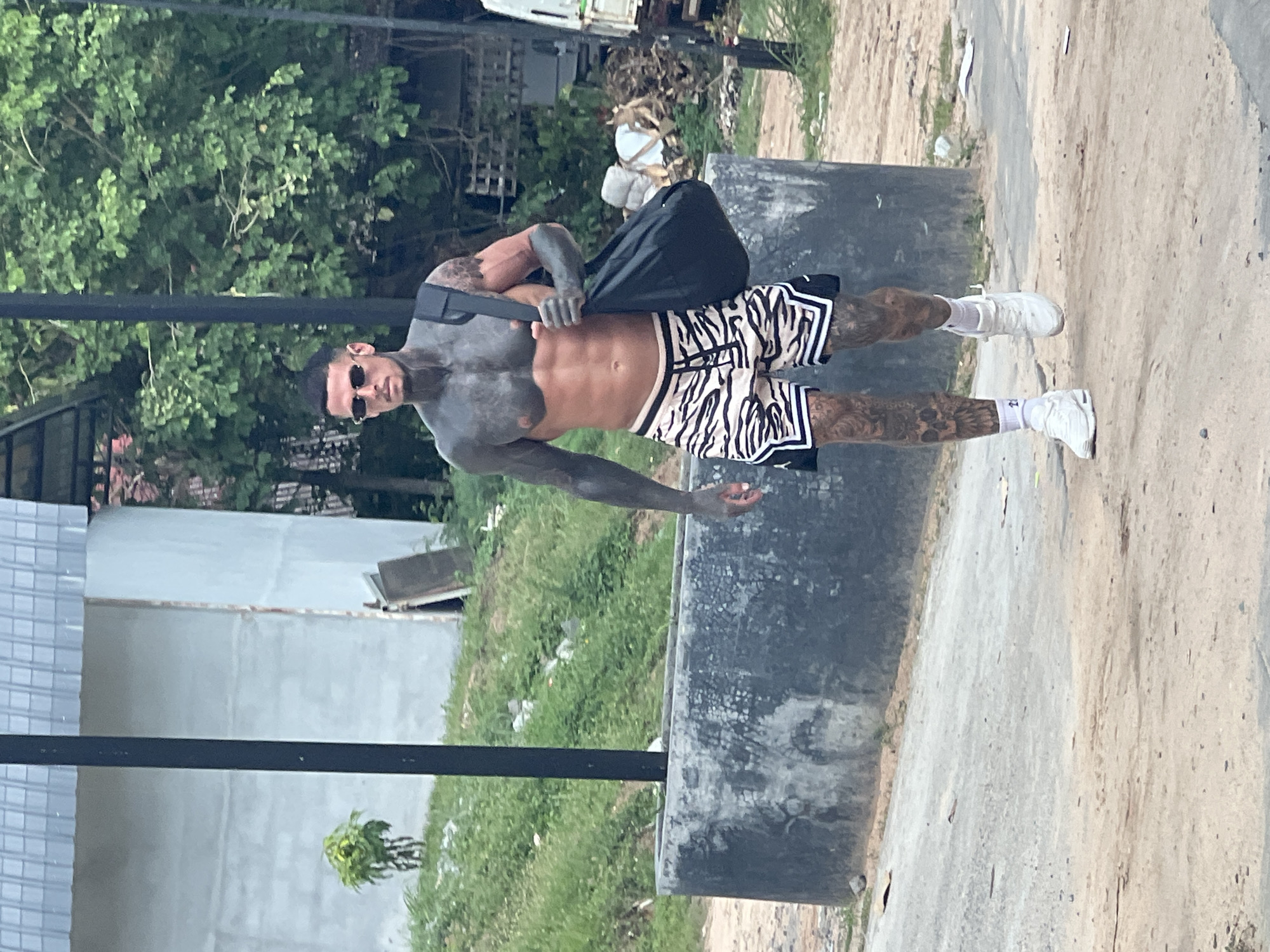 A heavily tattooed man walks along a dirt road in Ban Bang Rak, Thailand. He is shirtless, wearing white socks and sneakers, and shorts with a black and white zebra stripe pattern. He carries a black duffel bag over his right shoulder. The man is walking past a low concrete structure and a patch of overgrown grass. In the background, there are trees, buildings, and what appears to be construction materials.