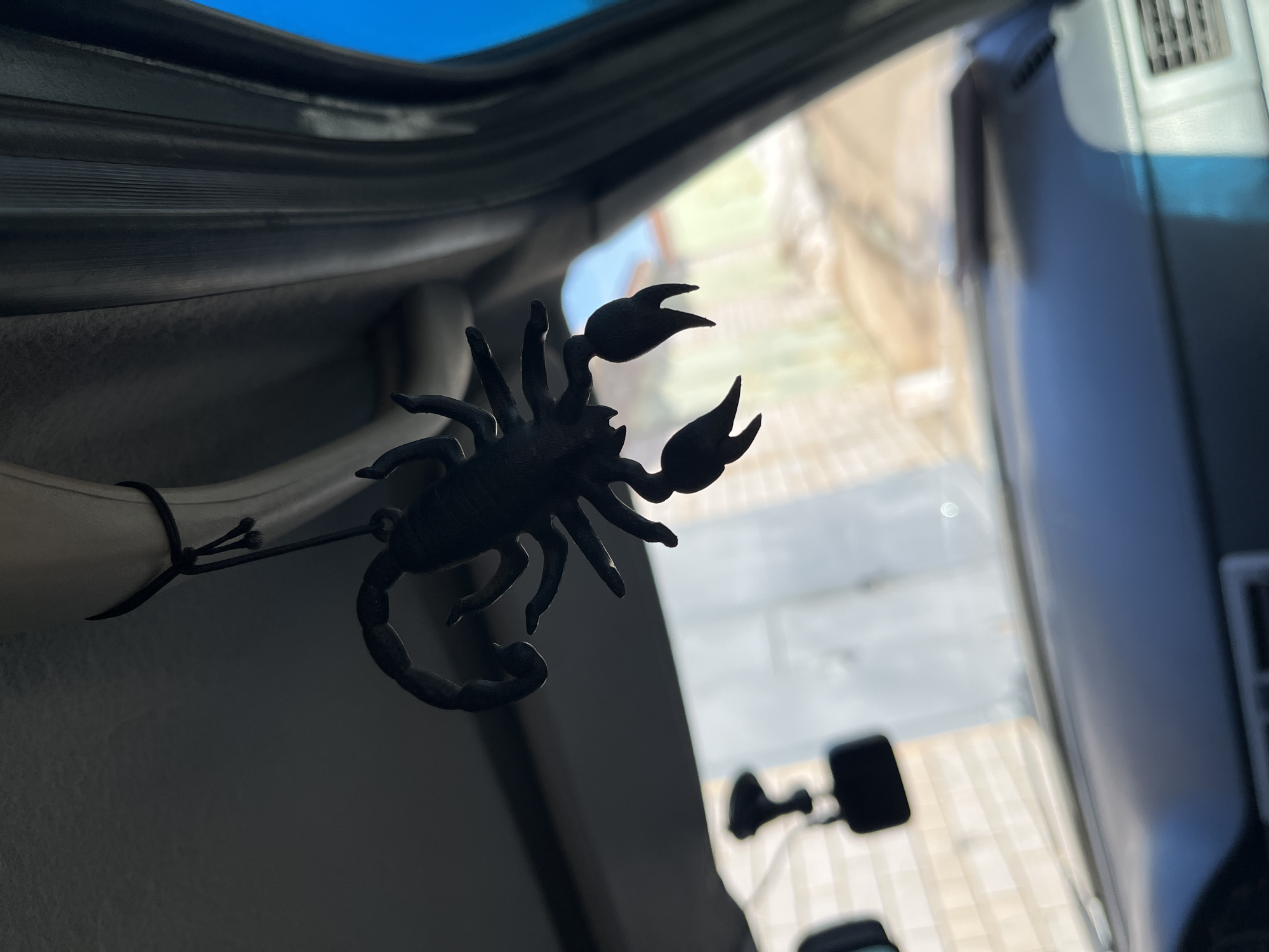 The image displays a close-up interior view from a vehicle, looking outward through the windshield. A black, stylized scorpion ornament hangs prominently from a black cord, tied around a light-colored interior fixture on the vehicle's headliner. The scorpion is silhouetted against the brighter background, with its tail curled and two prominent pincers visible.
To the right of the hanging scorpion, a dark, ribbed A-pillar or interior trim piece is visible. The vehicle's dashboard, dark blue or black, occupies the lower portion of the frame, with a grey air vent partially visible in the bottom right corner. A dashcam or similar device, black and rectangular, is partially visible hanging from the windshield in the upper-left background, obscured by the scorpion ornament.
The exterior environment, visible through the windshield, is out of focus. It shows a light-colored brick or textured building on the left, a blurred expanse of tan or light brown ground suggestive of a dirt path or unpaved area, and some indistinct lighter green/yellowish background elements. A patch of clear blue sky is visible through the far right side of the vehicle's window frame. The scene is brightly lit by natural sunlight from outside. There are no individuals or specific actions discernible. The setting is an outdoor, likely residential or industrial area, but a precise location within the city of Chaikovsky, Russia, cannot be determined from the visual evidence.
