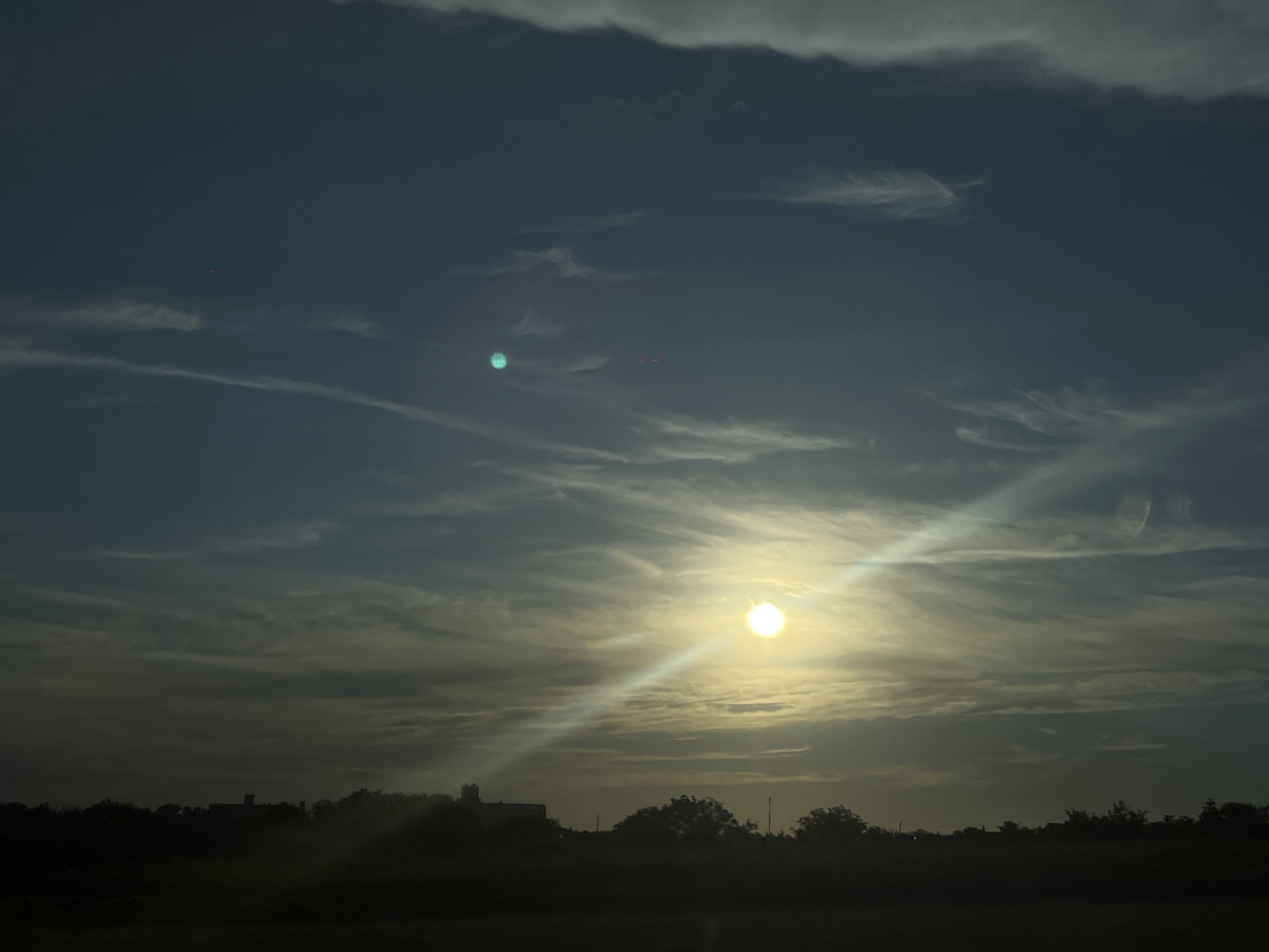 The image shows a silhouette of buildings and trees against a dusky sky. The sun is visible in the sky, casting a bright glare and a beam of light downwards. There are scattered clouds in the sky, with some appearing wispy and others more dense. The lower part of the image is dark, obscuring details of the ground and any potential figures. The scene appears to be outdoors, possibly at dusk or dawn, in a somewhat rural or suburban setting. The provided context mentions Novopokrovskaya, Russia, suggesting this is a landscape view from that location.