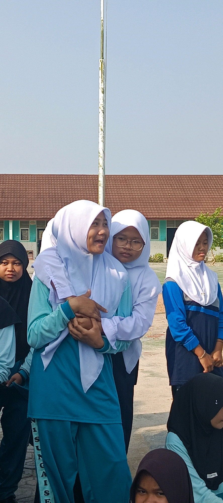 In Desa Sarang Mandi, Indonesia, a group of young females, most wearing hijabs, are standing outdoors in what appears to be a schoolyard. The background features a building with a brown tiled roof and light blue walls, and a tall, weathered flagpole.
The foreground is dominated by several individuals. On the left, a young woman in a black hijab and a light blue shirt is looking towards the viewer. Next to her, another young woman, also in a light blue shirt, is embraced from behind by a third young woman wearing a white hijab and a white top. The embracing woman has her arms around the other's shoulders and is holding her hands. A fourth young woman with glasses, wearing a white hijab and white top, stands slightly behind and to the right of the embracing pair, looking to her right. Further to the right, a fifth young woman in a white hijab and a blue and dark blue uniform is looking down, with her hands clasped in front of her. Partially visible at the bottom right are two other individuals wearing dark hijabs. One of the young women in the center, wearing a teal colored long-sleeved shirt and pants, has text vertically printed on the side of her left pant leg. The sky is clear and blue.
