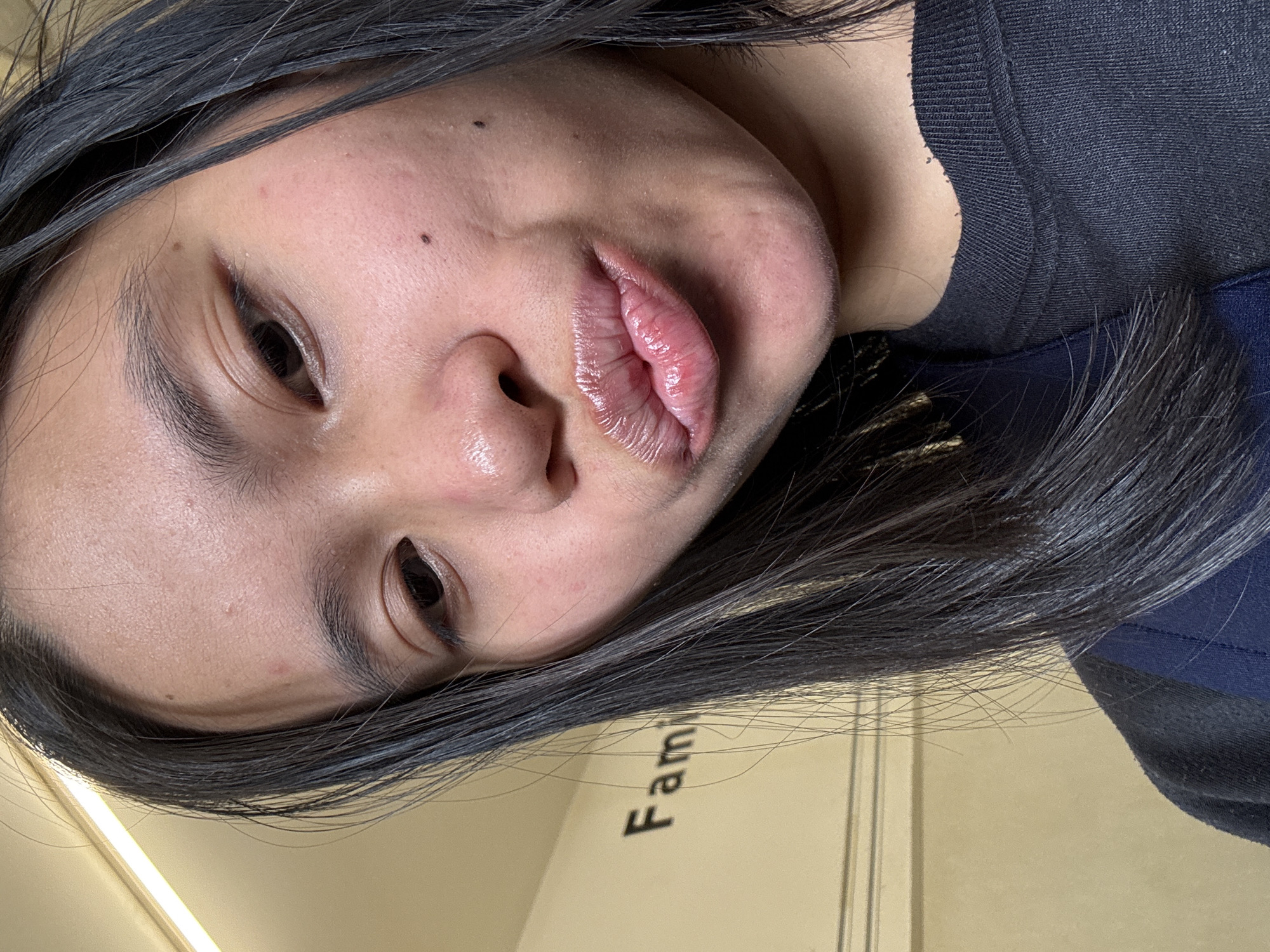 A close-up selfie of a young woman with long, dark hair, taken from the chest up. She is wearing a black t-shirt and her lips are pursed, wearing lip gloss. In the background, a wall is visible with the word "Family" written in black block letters. The lighting suggests an indoor setting, possibly a hallway or room. The location is specified as East Jakarta, Indonesia.