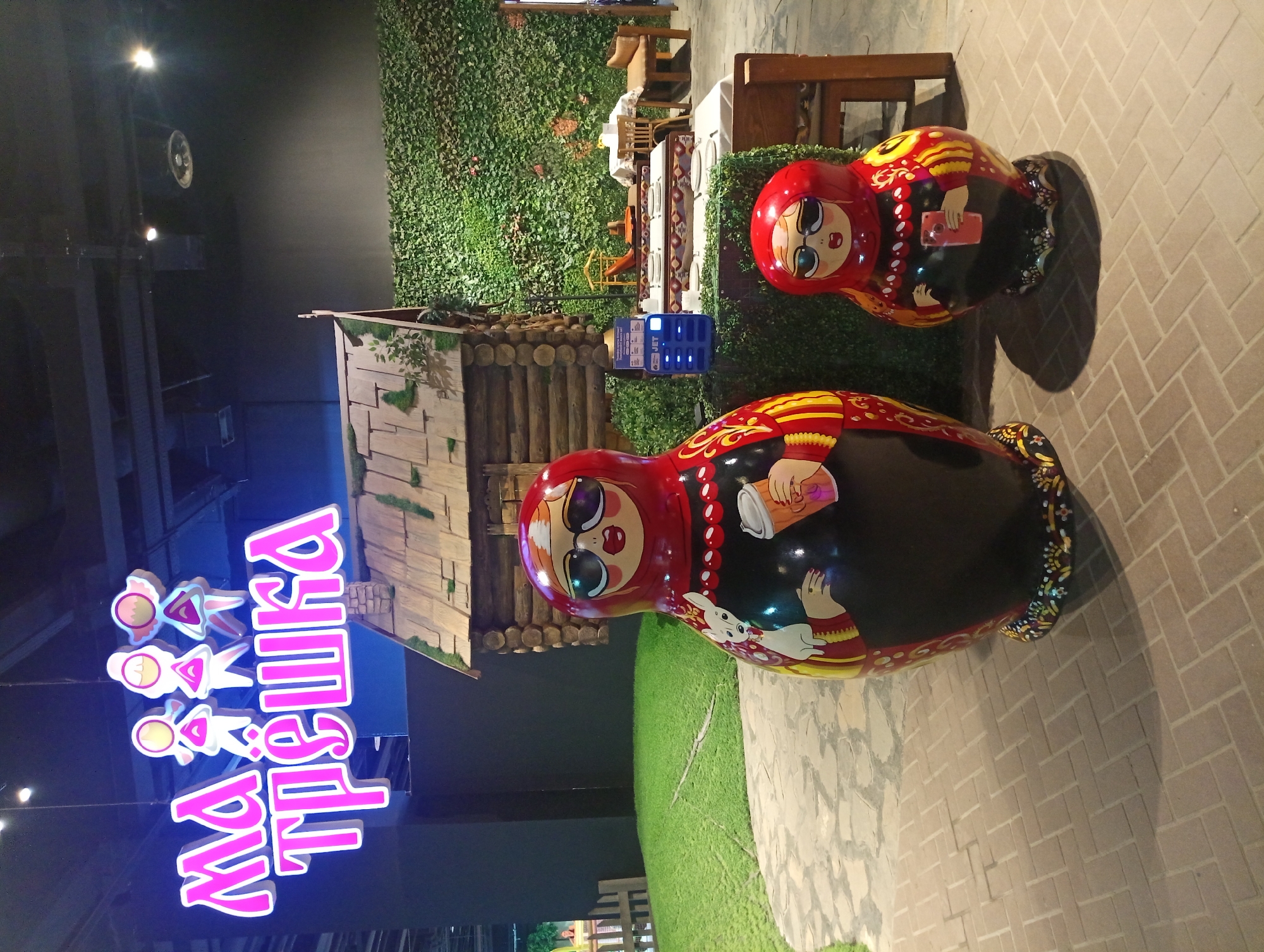 The image shows two large matryoshka dolls in the foreground, positioned on a paved area with a herringbone pattern. The dolls are brightly colored, depicting traditional Russian nesting dolls with modern accessories like sunglasses. The larger doll on the left is black and red, with a white cat painted on its chest and holding a coffee cup. The smaller doll on the right is also black and red, holding a pink smartphone.
In the background, there is a miniature log cabin with a shingled roof and moss accents, suggesting a rustic, possibly thematic, setting. Adjacent to the cabin is an area with artificial grass and a wooden fence, creating a stylized outdoor environment. Further back, a wall covered in dense green foliage serves as a backdrop. A set of tables and chairs are visible behind the dolls and cabin, arranged as if for dining, with white tablecloths and wooden furniture. A blue charging station with multiple ports is situated near the cabin.
Overhead, a neon sign in pink lettering reads "МАТРЁШКА," accompanied by three stylized figures. The overall impression is that of an indoor entertainment or dining venue with a Russian cultural theme, possibly a themed restaurant or play area. The lighting suggests an indoor environment, possibly a mall or a dedicated entertainment complex. The location is identified as Almaty, Kazakhstan.