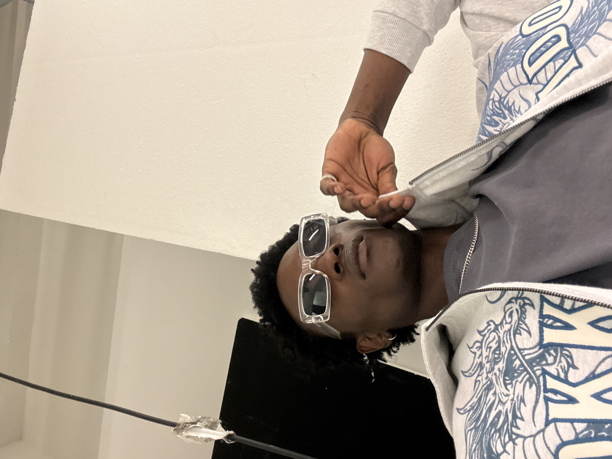 A man with dark skin and short, dark curly hair wears translucent sunglasses and a grey t-shirt under a white zippered jacket with a blue dragon design. He has a silver ring on his left hand and is holding the zipper pull of his jacket. The background includes a white wall and a black rectangular object. A black cord is visible in the lower left corner. The location cannot be determined from the image.