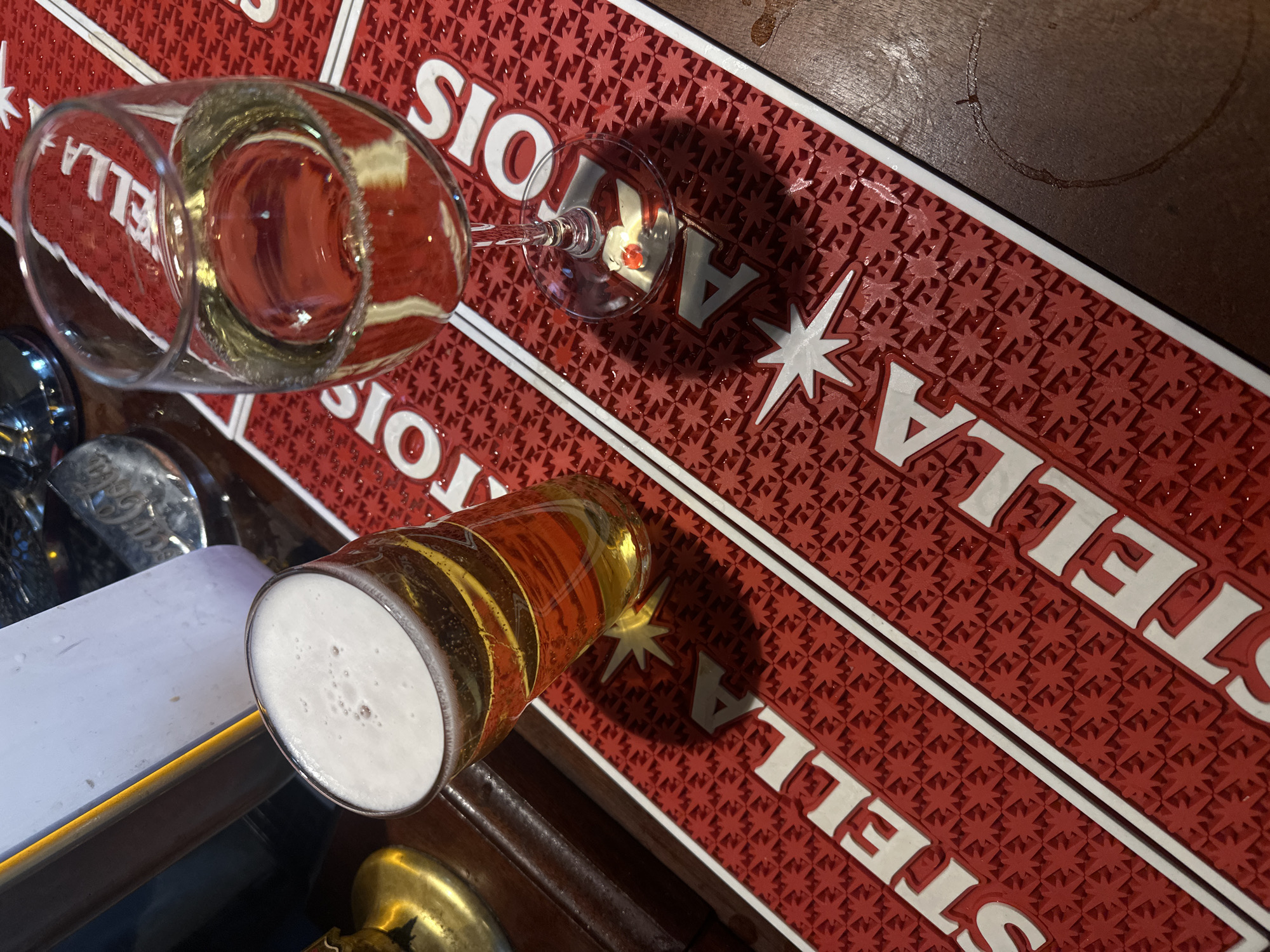 An overhead, slightly angled photograph captures two drinks resting on a red bar mat and a wooden surface in what appears to be a London bar or pub. The setting is likely indoors, possibly in the evening.
The main subjects are two glasses and the prominent red bar mat. One glass is a stemmed wine or champagne glass, filled with a clear, sparkling liquid, reflecting the red mat and its text. The other is a classic pint glass, containing a golden-amber beer with a substantial white foam head.
The drinks are situated on a distinctive red rubber bar mat that features a repetitive, raised star pattern. The word "STELLA" is clearly visible in white capital letters on the mat, and "ARTOIS" can also be seen, indicating the brand "Stella Artois." This branding is repeated across the mat, sometimes appearing inverted due to the angle. The mat is visibly wet in places, suggesting recent spillage or condensation from drinks. Adjacent to the mat, a dark wooden bar surface is visible, showing a circular wet ring mark where a glass once sat. Portions of metal bar fixtures and a white surface are also glimpsed in the foreground and background, hinting at typical bar equipment.
The scene suggests a relaxed social activity, likely drinking at a bar. The lighting casts subtle shadows, adding depth to the image. No people are visible.