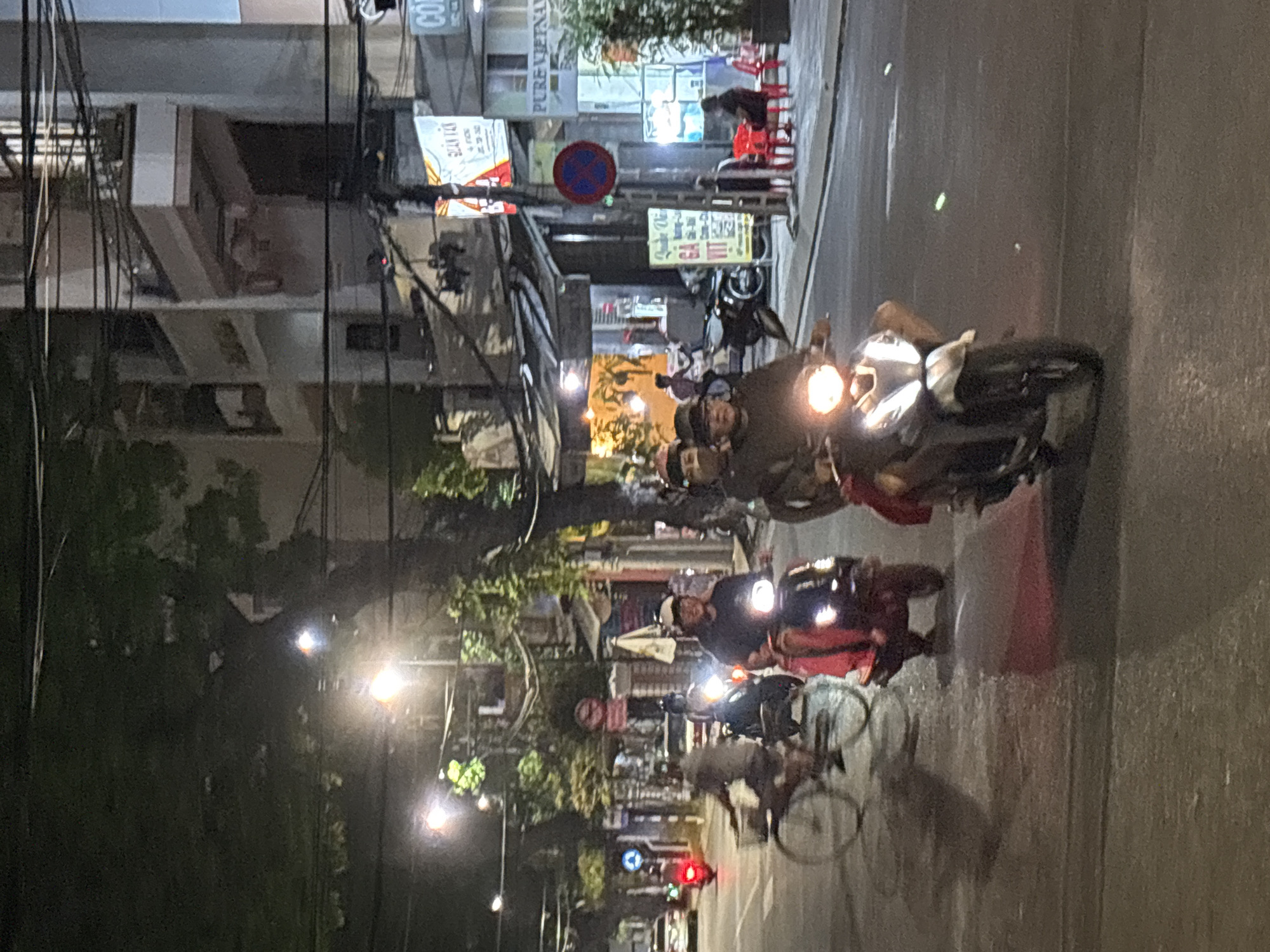 This is a nighttime street scene in Nha Trang, Vietnam. Several motorbikes and a bicycle are visible on a paved road, illuminated by streetlights and headlights. On the left, a person rides a bicycle with a basket. To their right, two people ride a red motorbike. In the foreground, two people ride a silver motorbike. Behind them, another person rides a motorbike. The background shows buildings, some with illuminated signs, and trees. The street is wet, indicating recent rain.