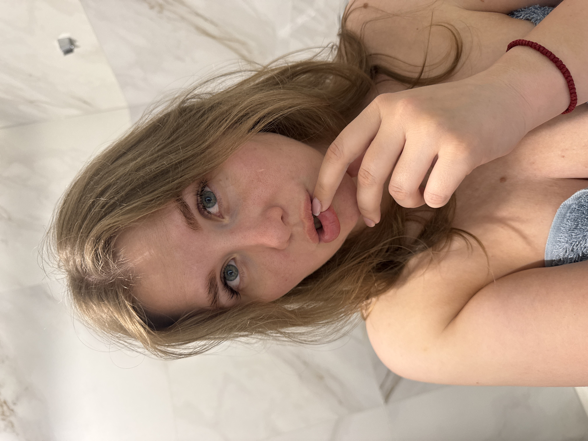 A young woman with light skin, blue eyes, and blonde hair is indoors in a bathroom with white marble tiles. She is partially wrapped in a light blue towel. Her right hand is near her mouth. She has a dark red bracelet on her left wrist. The location is in the city of Ферма, Russia.