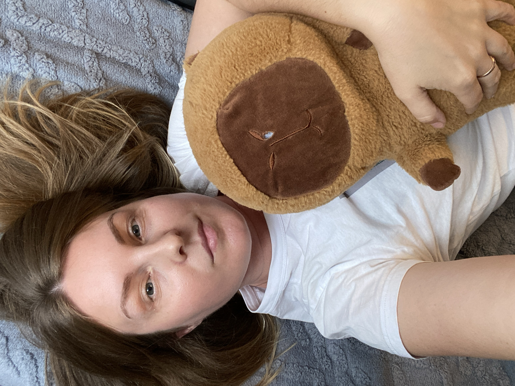 A woman is lying on a gray textured surface, possibly a bed or blanket. She is wearing a white t-shirt and is holding a brown stuffed animal with a darker brown face in her arms. She has fair skin, light brown hair, and is wearing a ring on her finger. The location is not identifiable as Noviy, Russia.