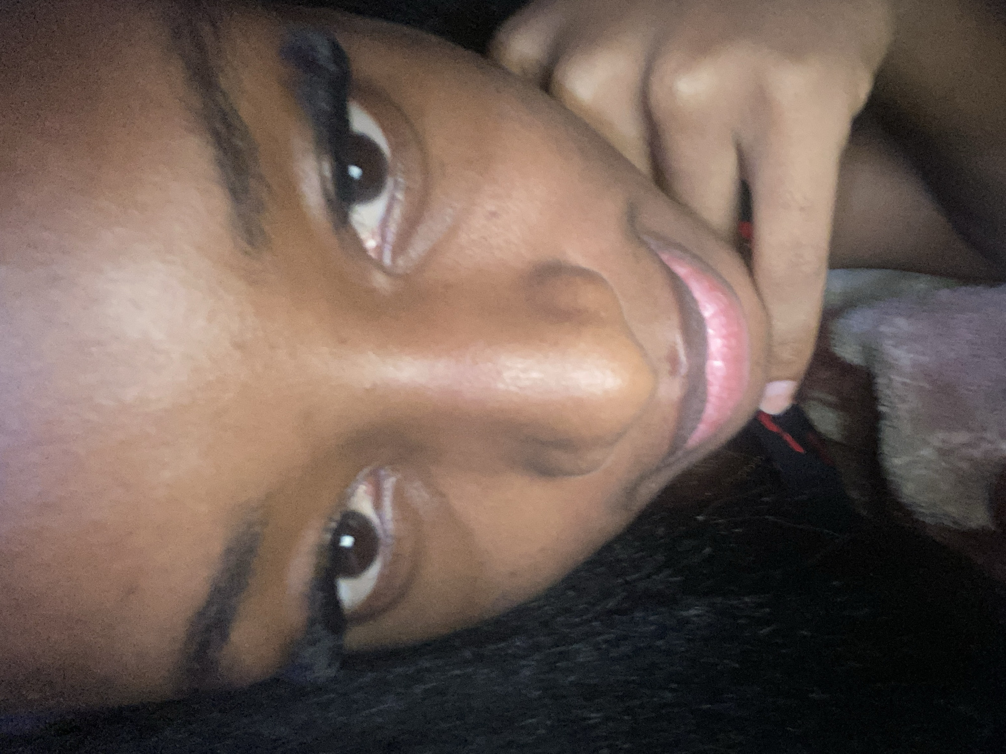 The image is a close-up, low-angle selfie of a young Black woman. She is looking directly at the camera with a neutral expression. Her eyes are wide open, and she is wearing false eyelashes. Her lips are slightly parted, revealing pinkish-red lipstick. Her skin is dark brown, and her hair is black and appears to be curly or wavy. A portion of her hand is visible near her chin, and she is wearing a dark-colored top with a thin red stripe. The background is dark and out of focus, suggesting an indoor setting or low-light conditions. There is no identifiable information in the image to indicate a specific location within Jos, Nigeria.
