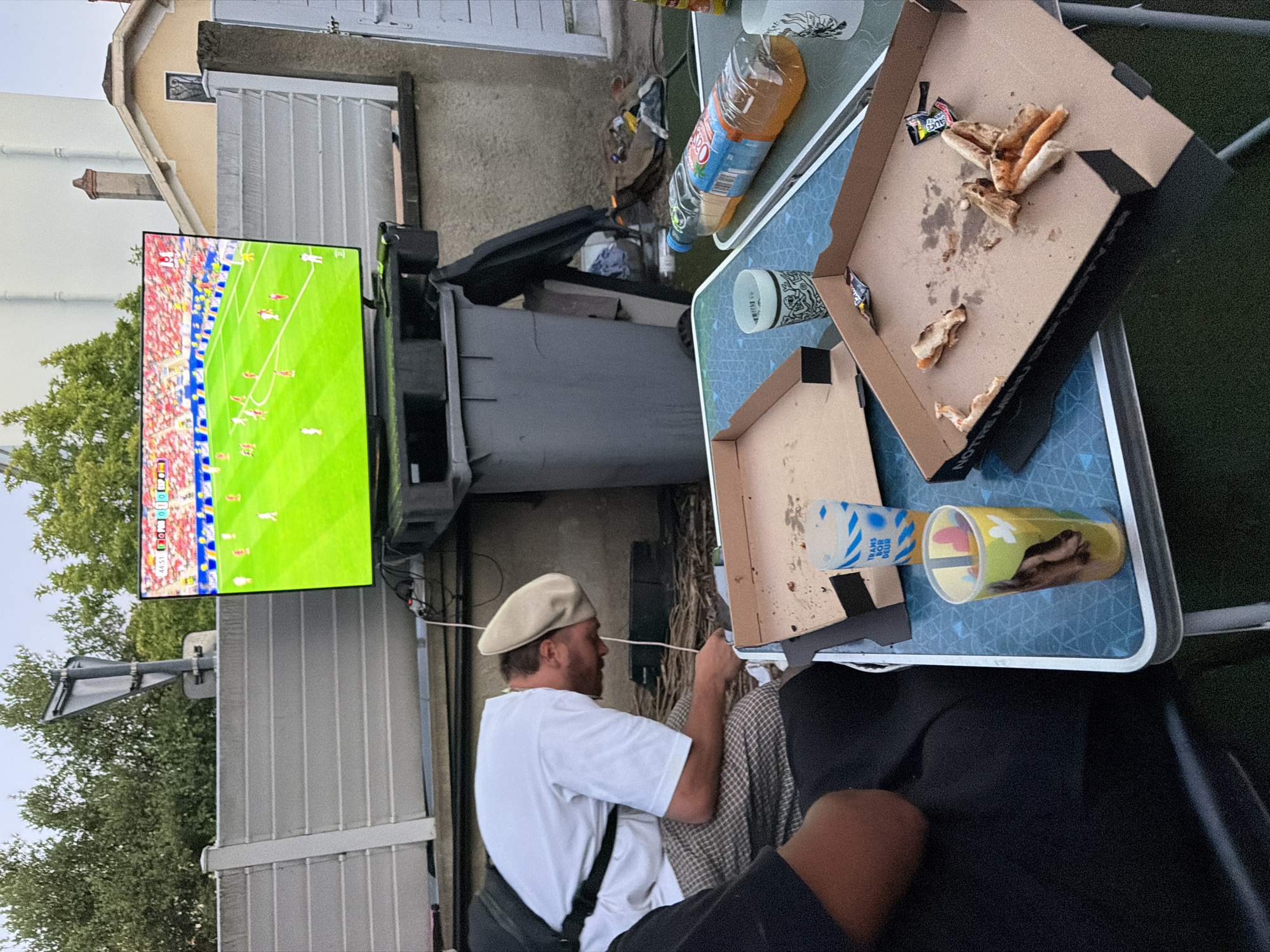 In Villeurbanne, France, an outdoor scene unfolds with a flat-screen television displaying a soccer match. A man, wearing a white polo shirt and a beige cap, is seated and looking down, appearing to be engaged with something on his lap. He is positioned in front of a table laden with remnants of a meal, including empty pizza boxes and disposable cups. One cup bears the text "TRANS BORDEUR." A large plastic trash bin is visible in the background, alongside a fence and some vegetation. The overall setting suggests a casual gathering, possibly in a backyard or patio area, during daylight hours.