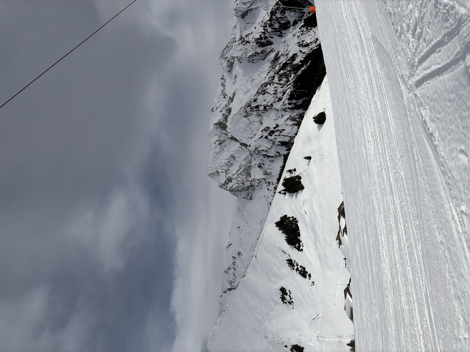 This image depicts a snow-covered alpine landscape in the city of Manigod, France, under an overcast sky. The foreground features a wide, groomed ski piste, exhibiting parallel lines from grooming machinery and distinct ski tracks in the snow. In the midground, a snow-covered hillside is visible, dotted with clusters of dark evergreen trees. Several buildings with dark, pitched roofs are partially obscured by snow at the base of this hillside. Farther up the slope, subtle ski lift infrastructure is discernable. The background is dominated by rugged, snow-capped mountain peaks with exposed rock formations. A dark, thin cable, consistent with a ski lift, stretches diagonally from the upper right corner of the frame downwards. A small, orange, cylindrical marker is partially visible on the right edge of the groomed slope. No individuals or animals are present within the scene.