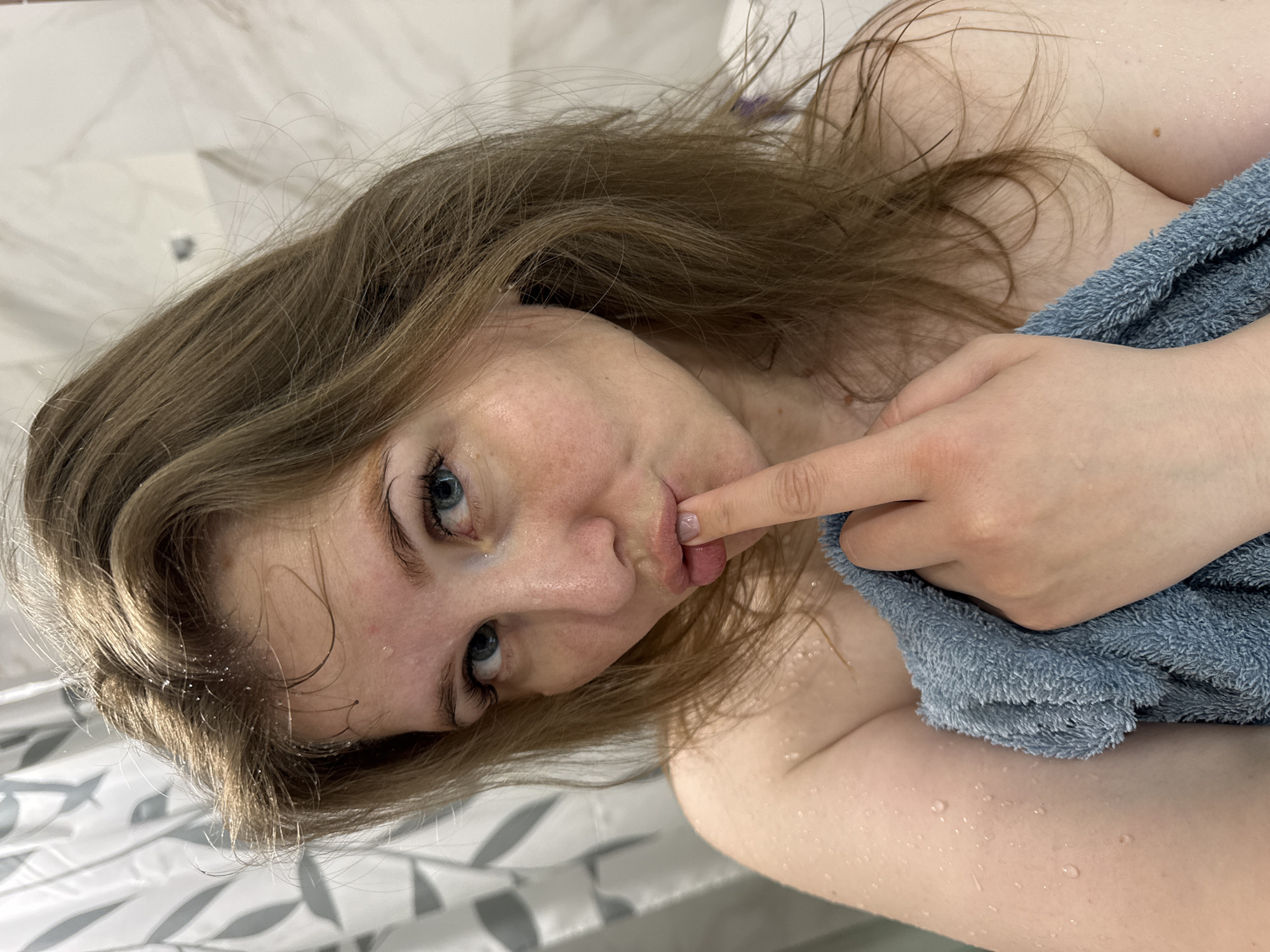 A young woman with light brown hair and striking blue eyes is depicted in a close-up, angled shot. Her hair is wet, and her skin, chest, and arms are covered with water droplets, suggesting she has recently showered or bathed. She is partially wrapped in a soft, light blue towel, which is visible around her upper body. Her right hand is raised, with her index finger gently touching her lips, creating a playful or suggestive expression. She is looking directly towards the viewer. The background features light-colored, possibly white or off-white, tiled walls, consistent with a bathroom or shower environment. No text is visible in the image. The setting is indoors, with the location context being Gornyi (Dvurechenskoe S/P), Russia.
