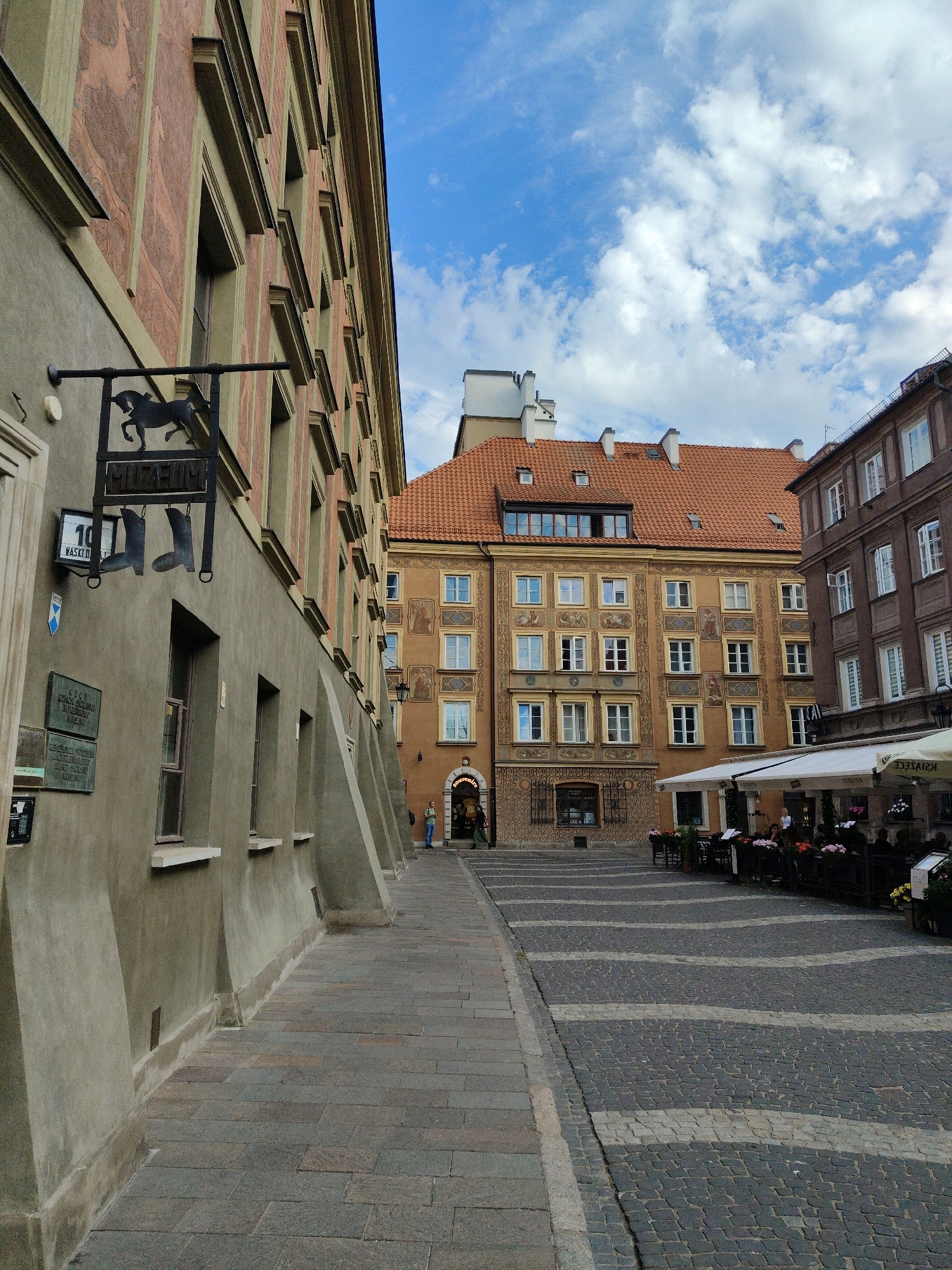 The image captures a view of the Old Town Market Square in Warsaw, Poland. The foreground is dominated by a narrow, paved alleyway to the left, with the textured facade of a building displaying a horse and boot emblem above a sign. To the right, a wider, cobblestone plaza is laid out with a distinctive wavy pattern of lighter stones. This plaza is lined with tables and chairs belonging to an outdoor cafe with a white awning. In the background, several historic buildings with terracotta roofs are visible. One building in the center features ornate yellow facades with decorative paintings between windows, while the building to its right is a darker brown with multiple windows. A few people are present, including one person standing near the cafe and another walking in the background on the right. The sky above is blue with scattered white clouds.